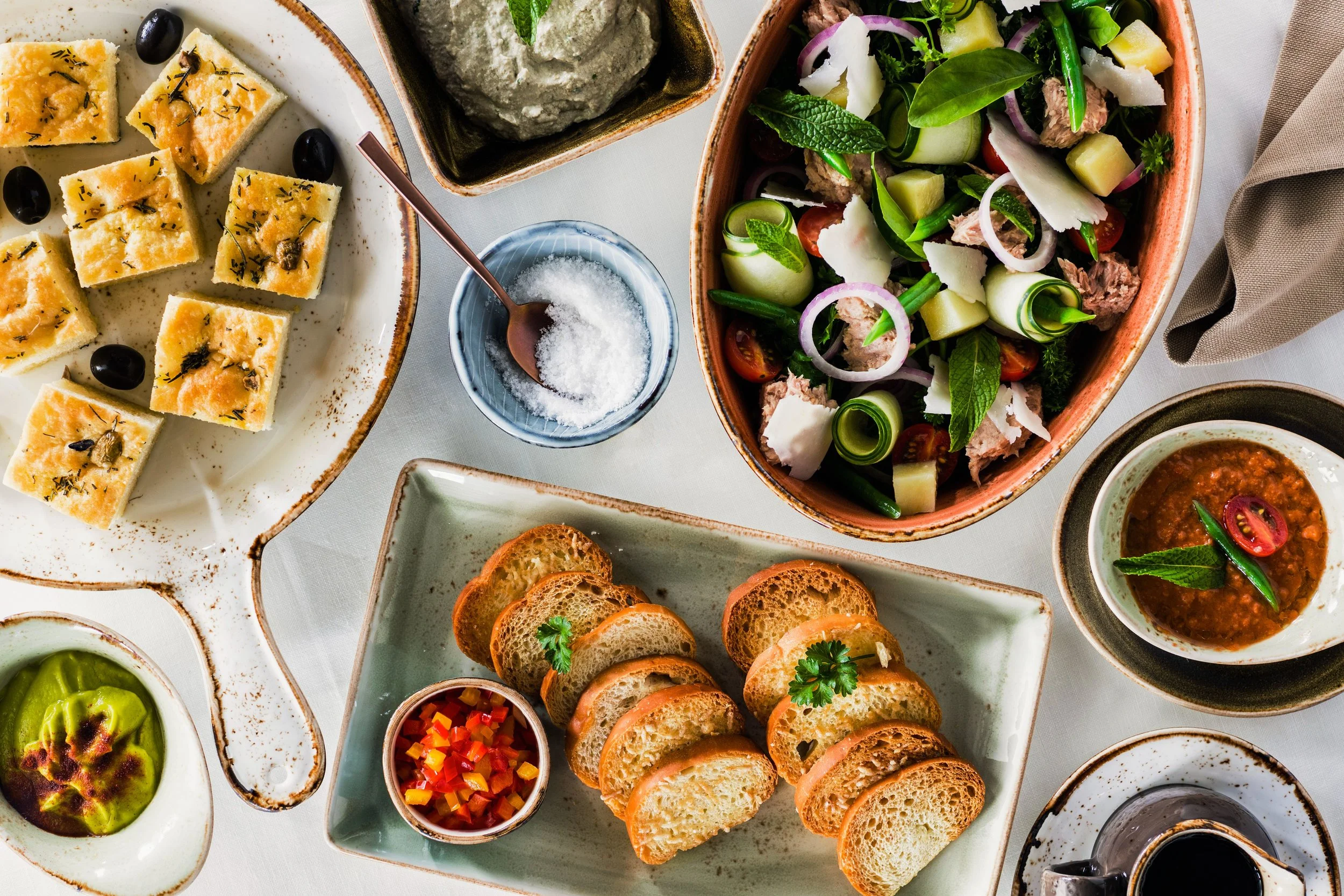 An assortment of Mediterranean dishes on a white table, including sliced bread, stuffed grape leaves, vegetable dips, salad with cucumber, tomatoes, onion, and herbs, and small bowls of various toppings and sauces.