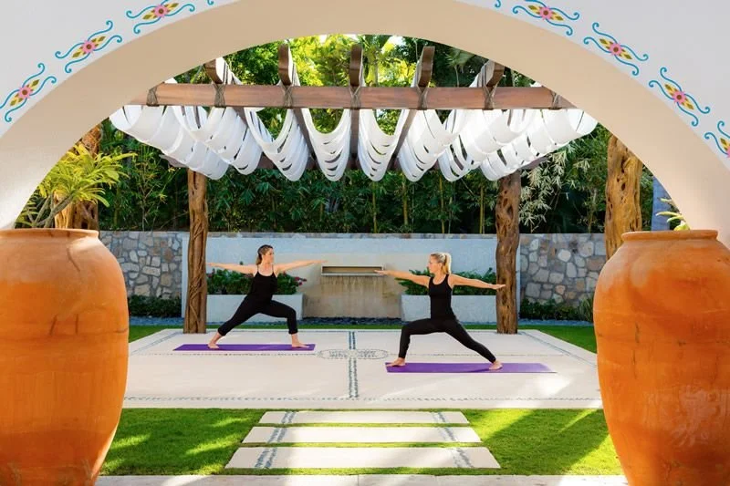 Two women practicing yoga outdoors on purple mats under a wooden pergola, surrounded by large clay pots and green trees.