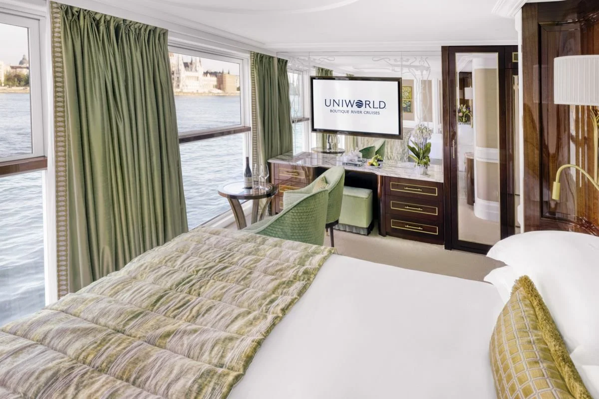 Luxury cruise ship room with large windows overlooking a river and city, green curtains, a bed with a patterned quilt and a pillow, a small table with a bottle and glasses, a green chair, a TV screen displaying 'UNIWORLD BOUTIQUE RIVER CRUISES', marble desk with plants, and a closet with glass doors.