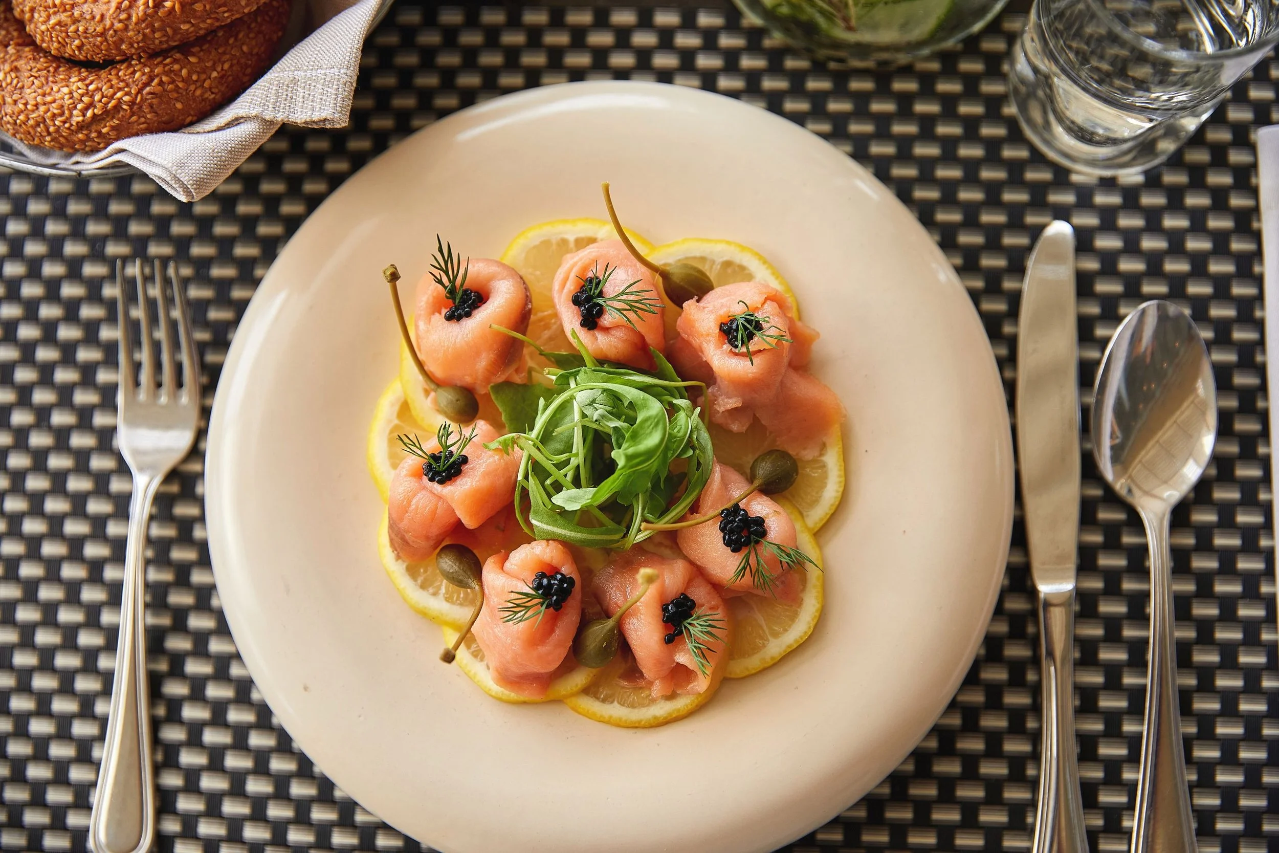 A plate of smoked salmon with lemon slices, capers, black caviar, and fresh herbs, served on a beige plate with silver cutlery and a glass of water in the background.