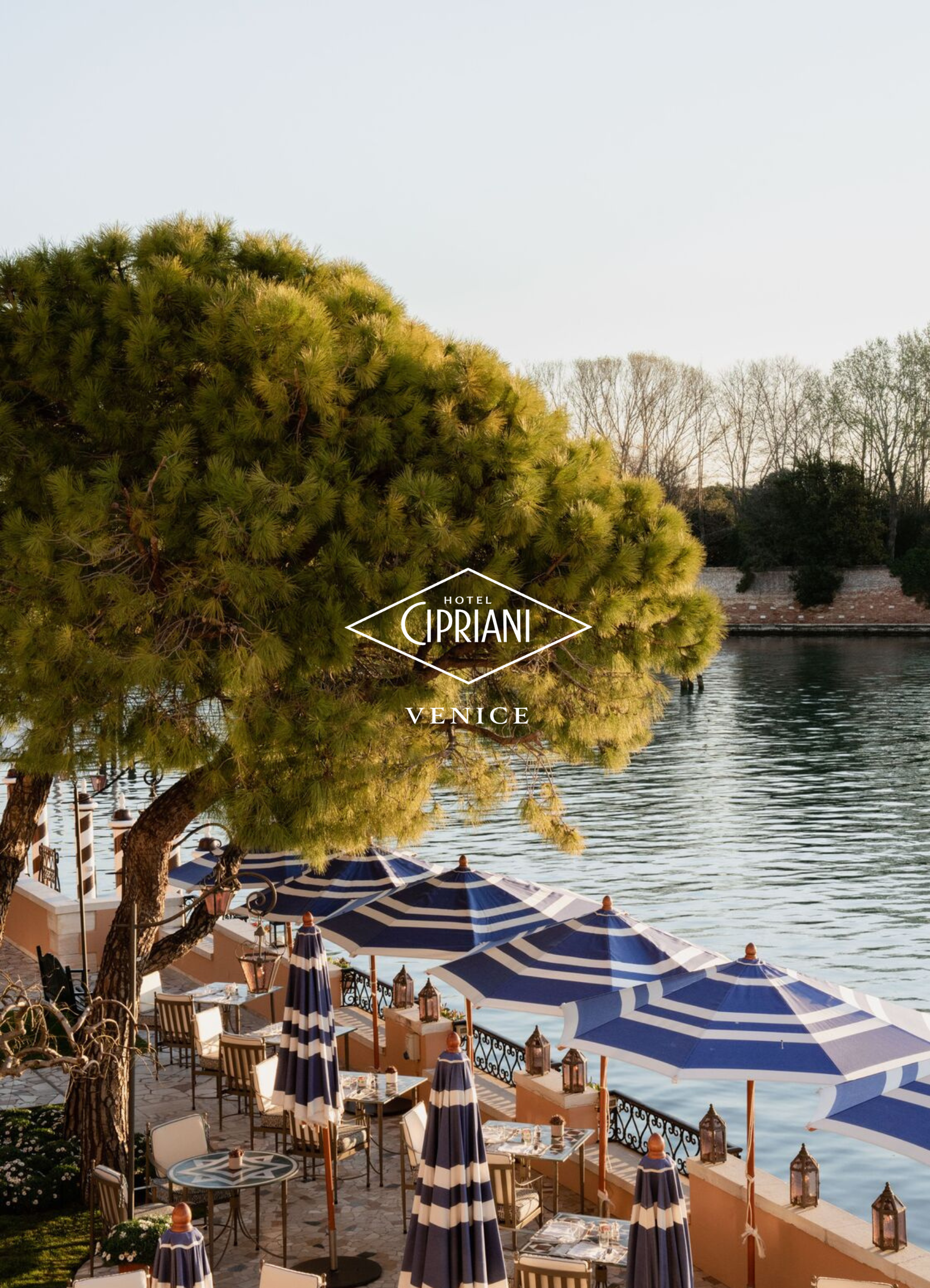 Outdoor dining area at Hotel Cipriani in Venice with tables, chairs, and blue-and-white striped umbrellas near a waterway, framed by green trees.