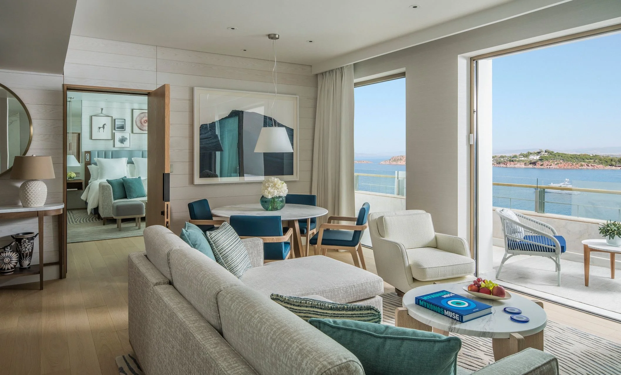 Living room with large windows overlooking water, furnished with beige sofa, armchairs, round coffee table with books and fruit, and a dining area visible through an open door, with bedroom beyond.