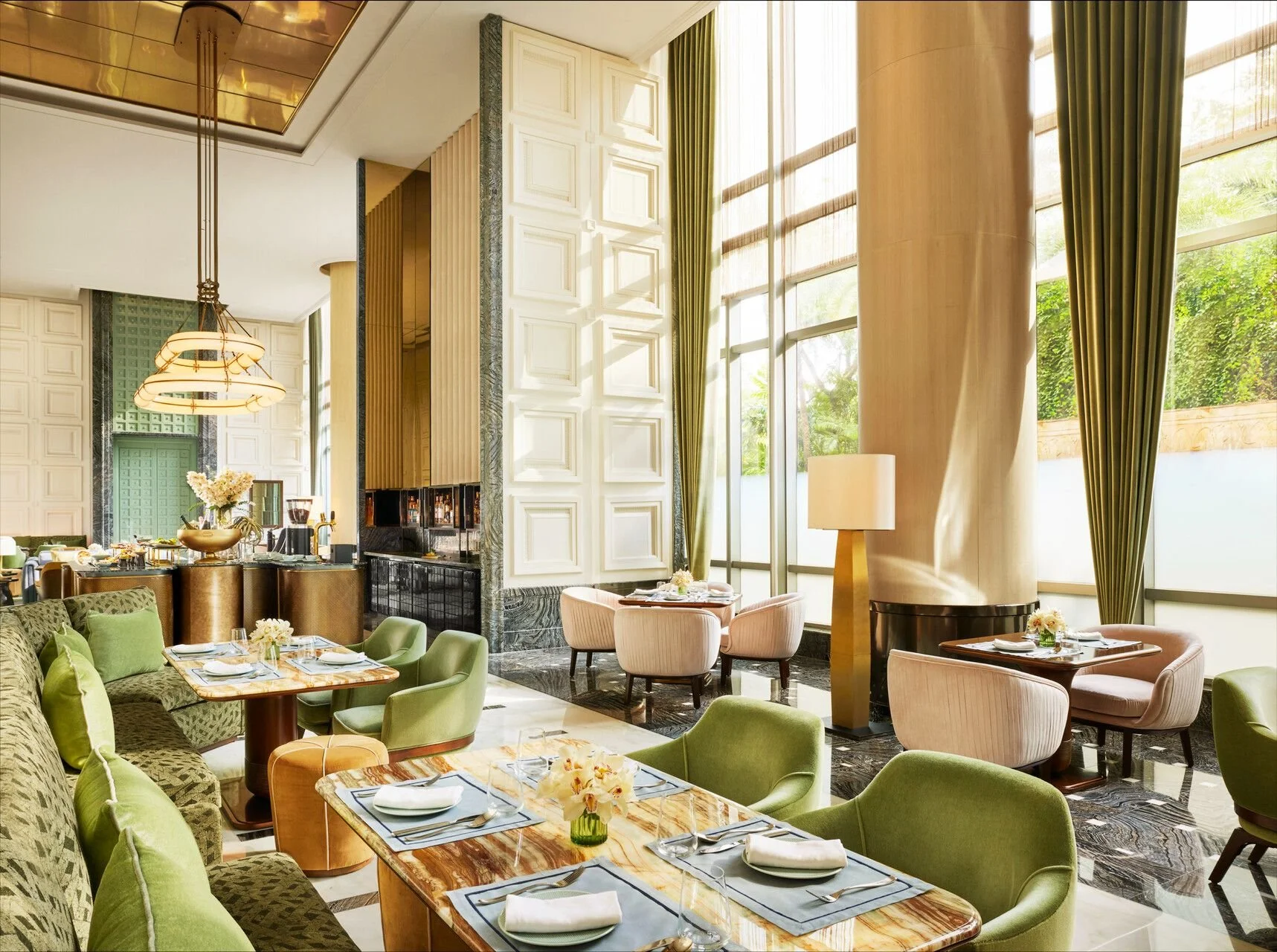 Elegant restaurant interior with green and beige seating, marble-topped tables set with napkins, plates, and flowers, large windows with green curtains, tall beige columns, and a high ceiling with gold and white accents.