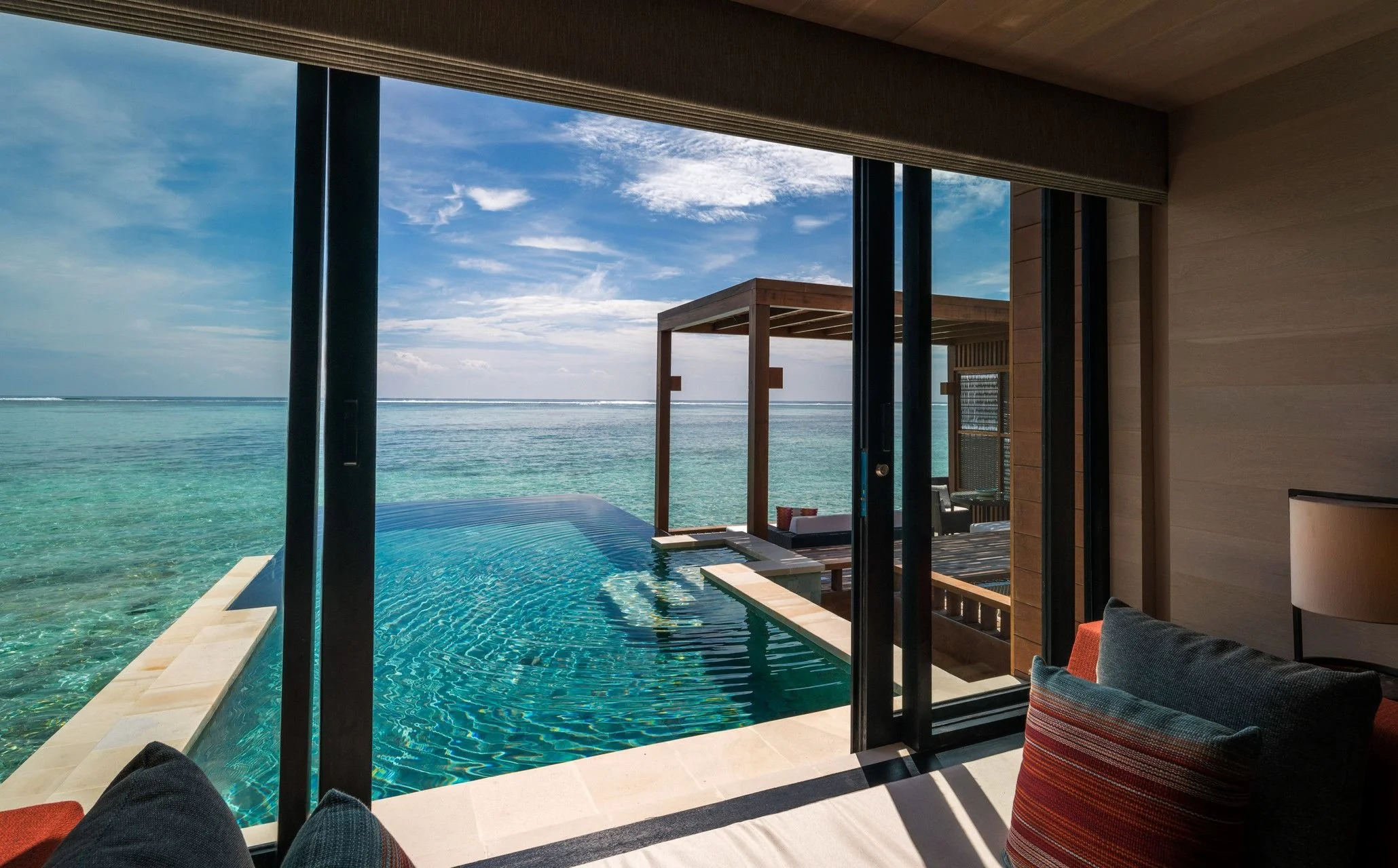 View from inside a modern beachfront villa showcasing a private infinity pool overlooking the ocean, with a terrace and blue sky with scattered clouds.