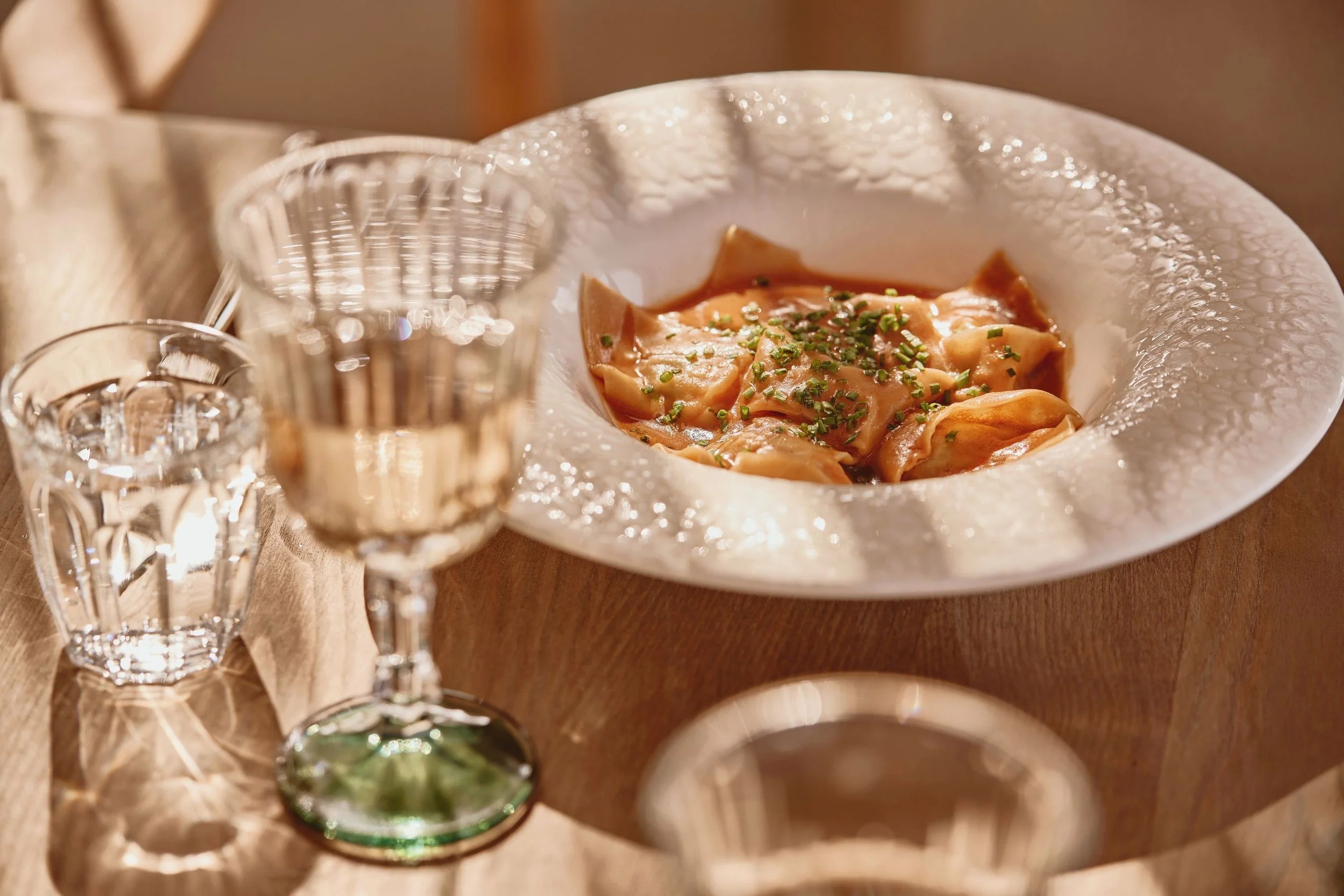 A white bowl with textured surface containing pasta with sauce and chopped herbs, set on a wooden table with multiple empty champagne glasses.