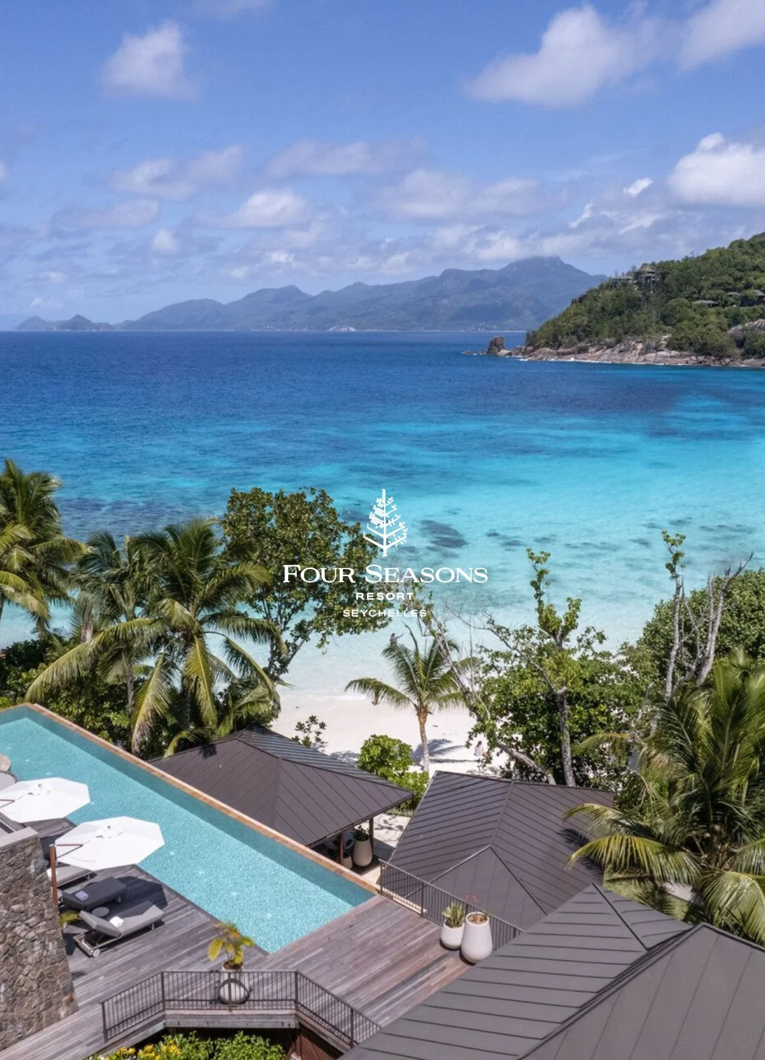 An aerial view of a resort with a swimming pool, surrounded by lounge chairs and umbrellas, trees, and buildings with metal roofs. In the background, blue ocean waters and a mountainous coastline are visible under a partly cloudy sky. The logo for Four Seasons Resort Seychelles is overlaid on the image.