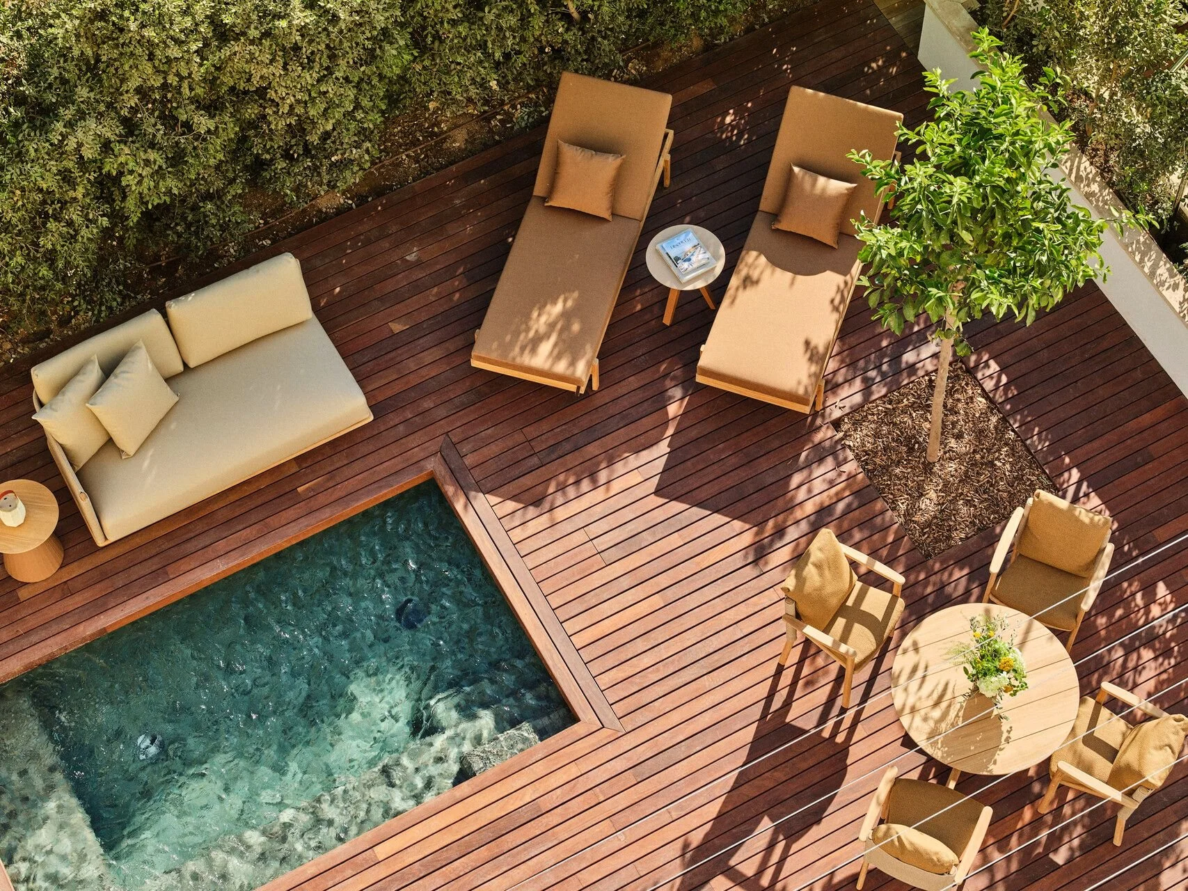 An aerial view of a wooden deck with outdoor furniture, including a sofa, sun loungers, a round table with chairs, and a small side table next to a pool and surrounding greenery.