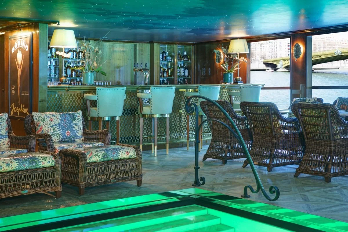 Indoor lounge area with wicker chairs, a bar in the background with liquor bottles, large windows overlooking a river and cityscape, and a ceiling painted to resemble the night sky with stars.
