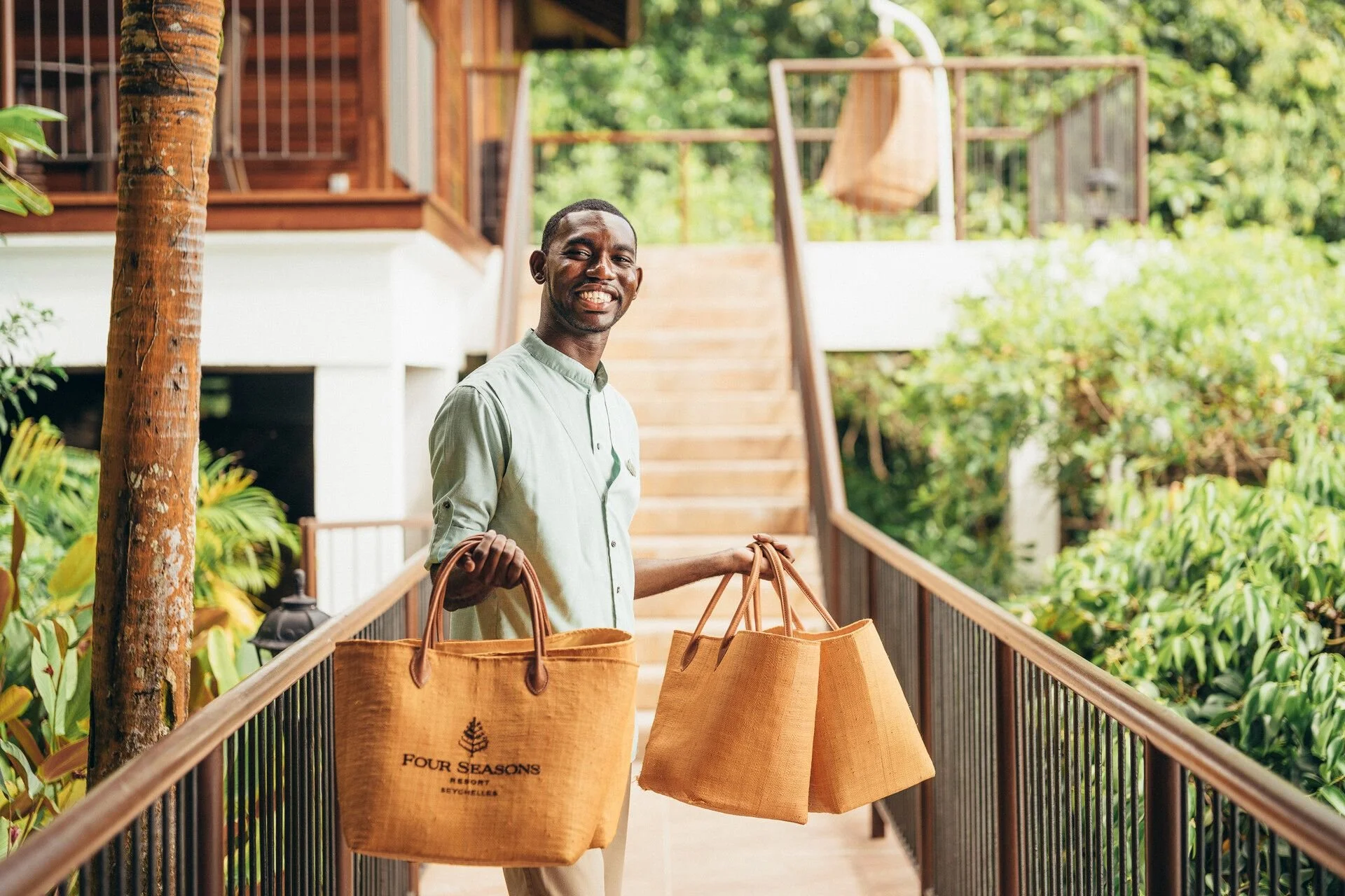 Smiling man on a wooden walkway holding reusable shopping bags with "Four Seasons Resort Seychelles" logo, surrounded by lush greenery.