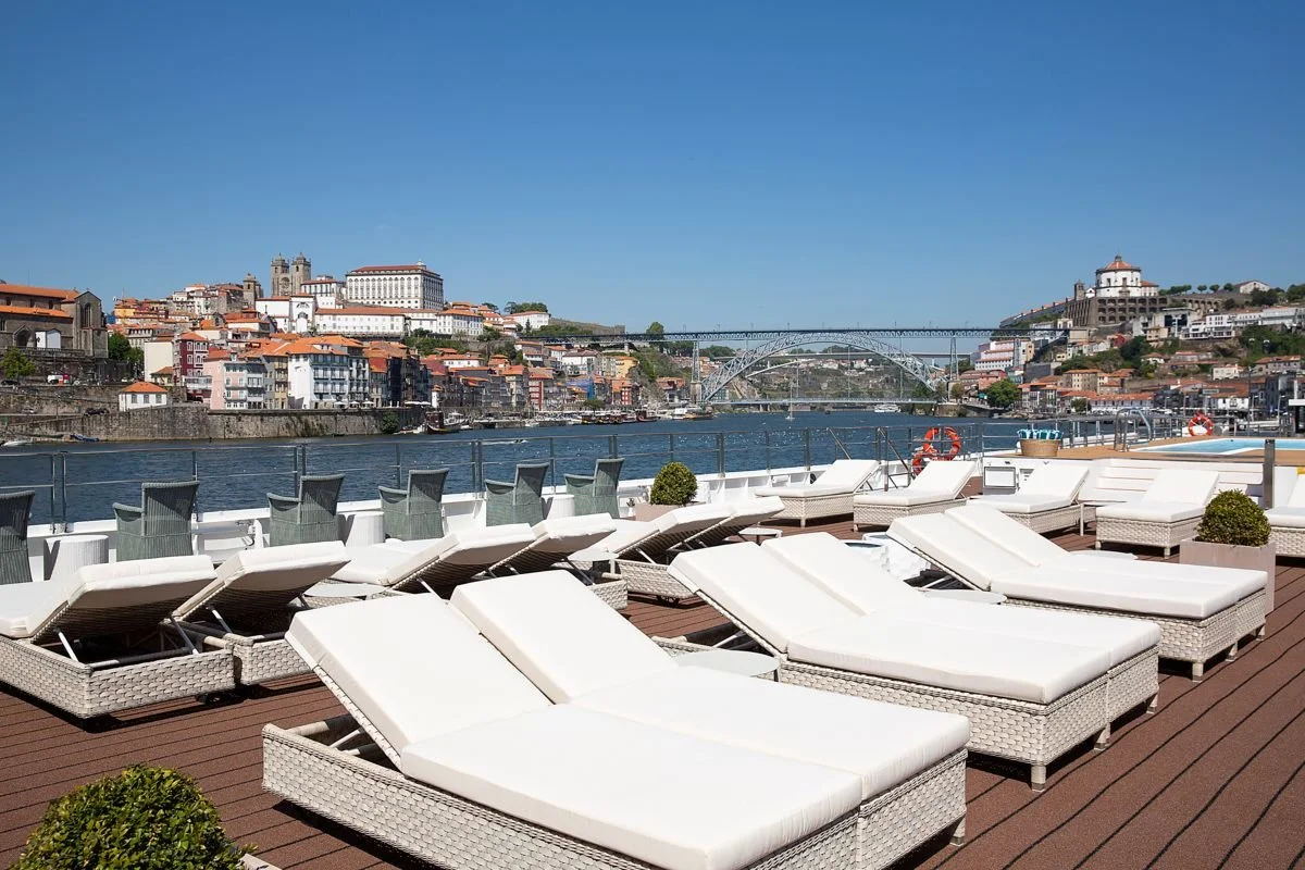 Luxury cruise ship deck with white lounge chairs and view of the city of Porto, Portugal, including Dom Luís I Bridge and Ribeira district along the Douro River.