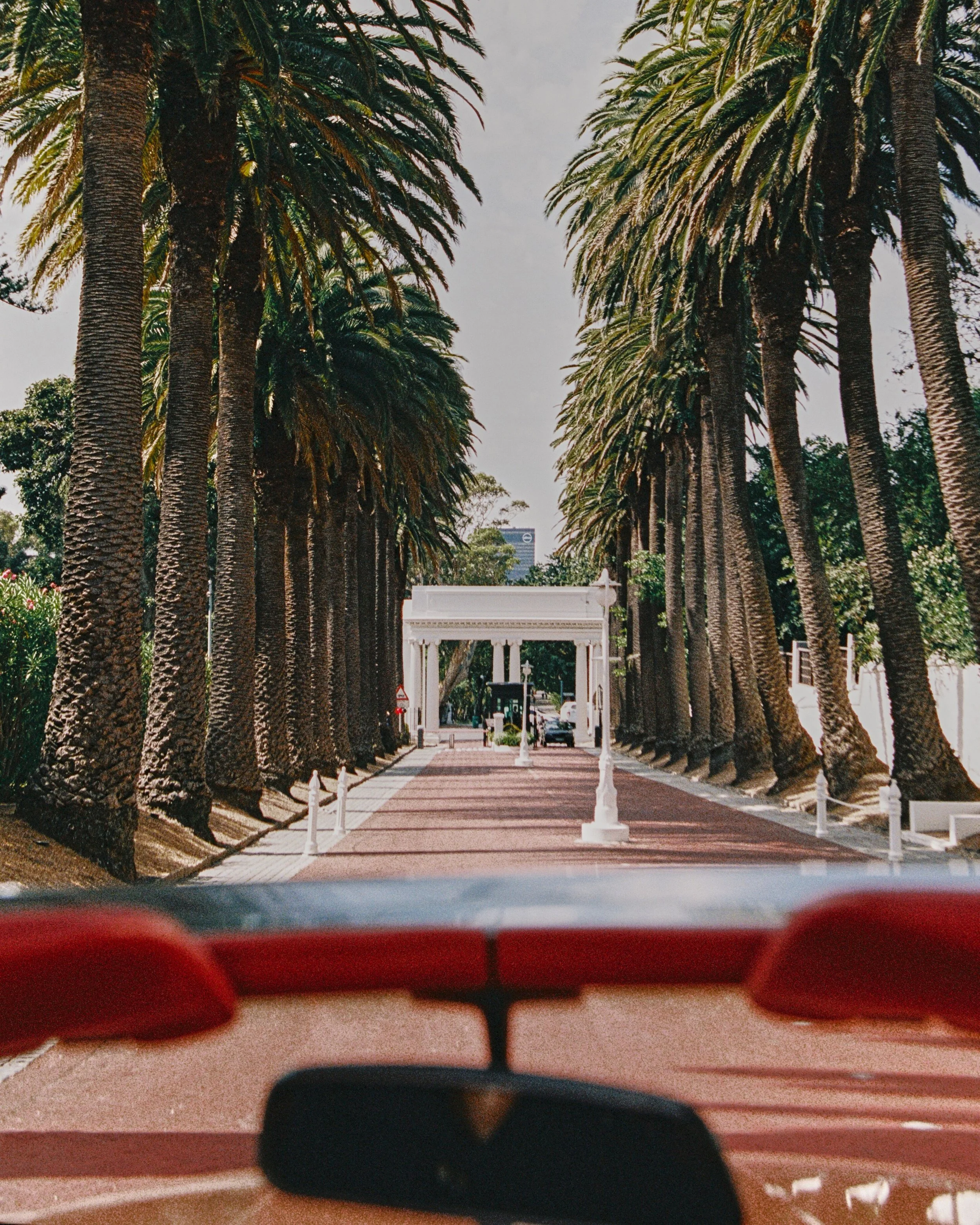 View through a car's windshield showing tall palm trees lining a street with a white archway and tiki statues in the distance, under a cloudy sky.