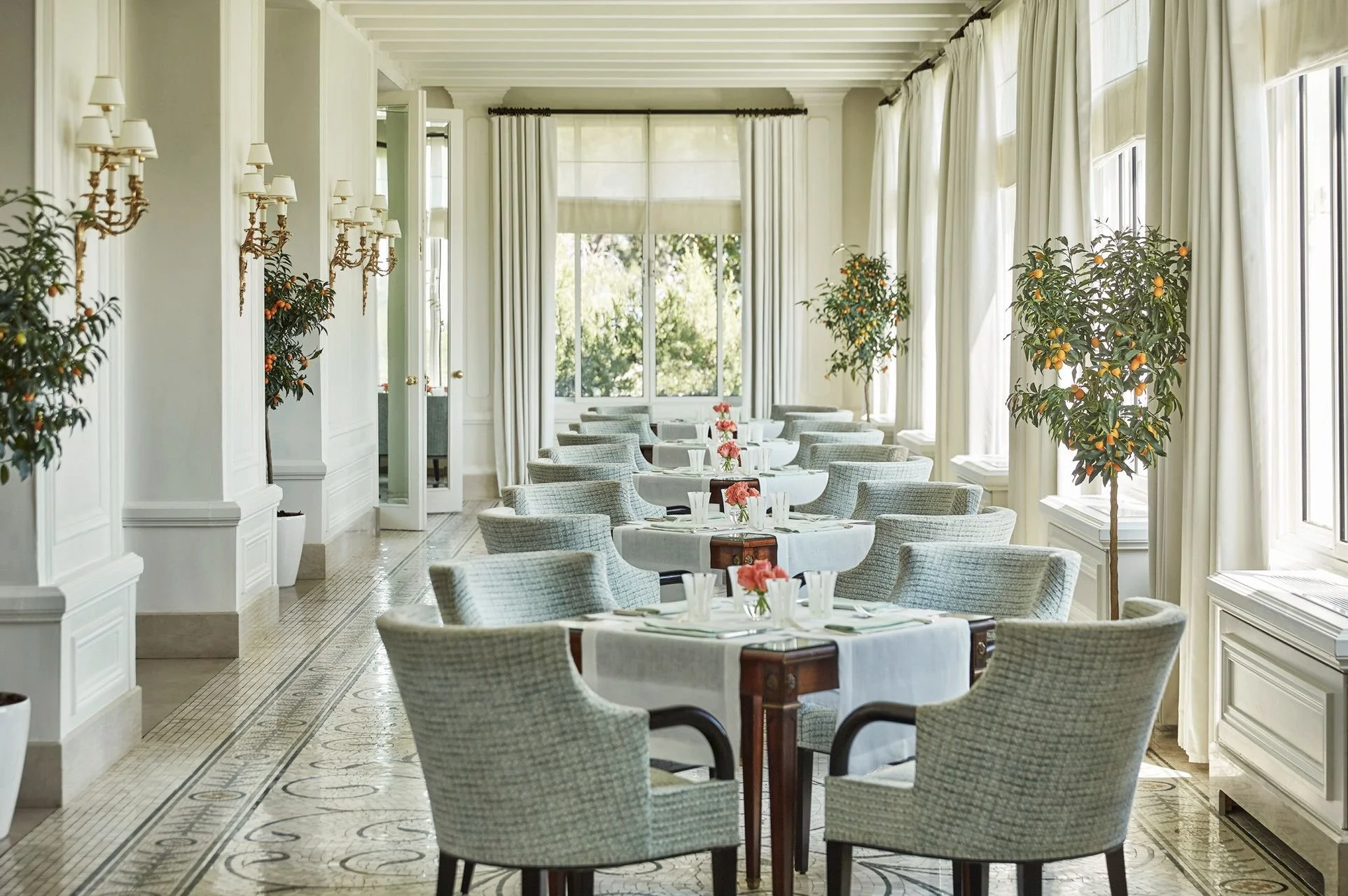 Elegant dining room with round tables, white tablecloths, pink floral centerpieces, gray upholstered chairs, large windows with white curtains, potted orange trees, and wall-mounted brass candle sconces.