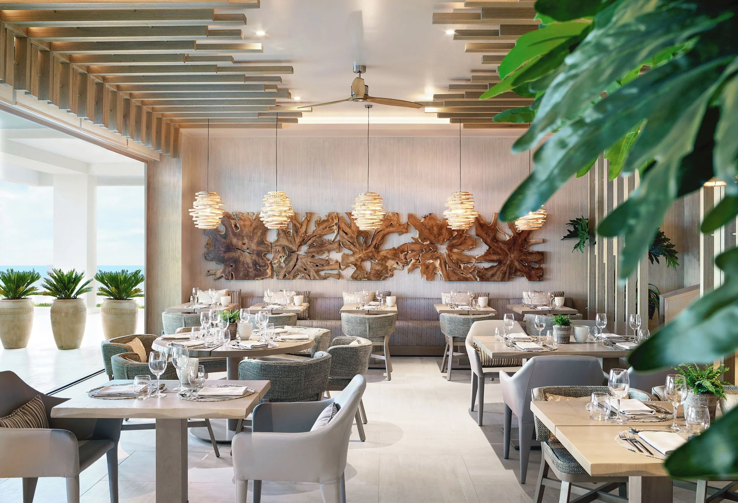 Elegant restaurant dining area with round tables, upholstered chairs, and decorative wood wall art, illuminated by pendant lights and natural sunlight, surrounded by green plants.