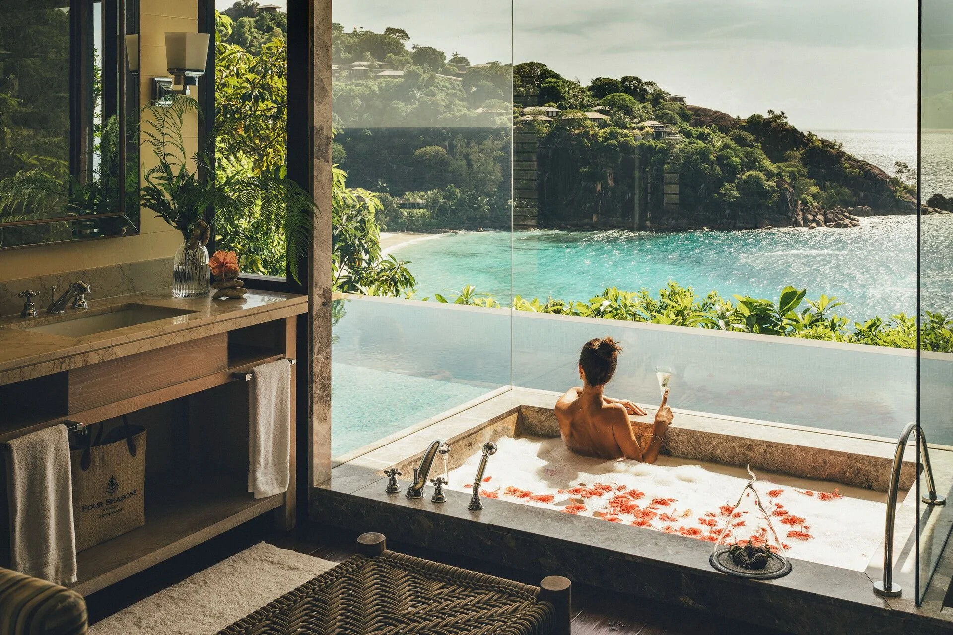 A woman relaxing in a bathtub on a balcony overlooking ocean and green hillside, holding a glass of wine, with tropical plants and a bathroom sink nearby.