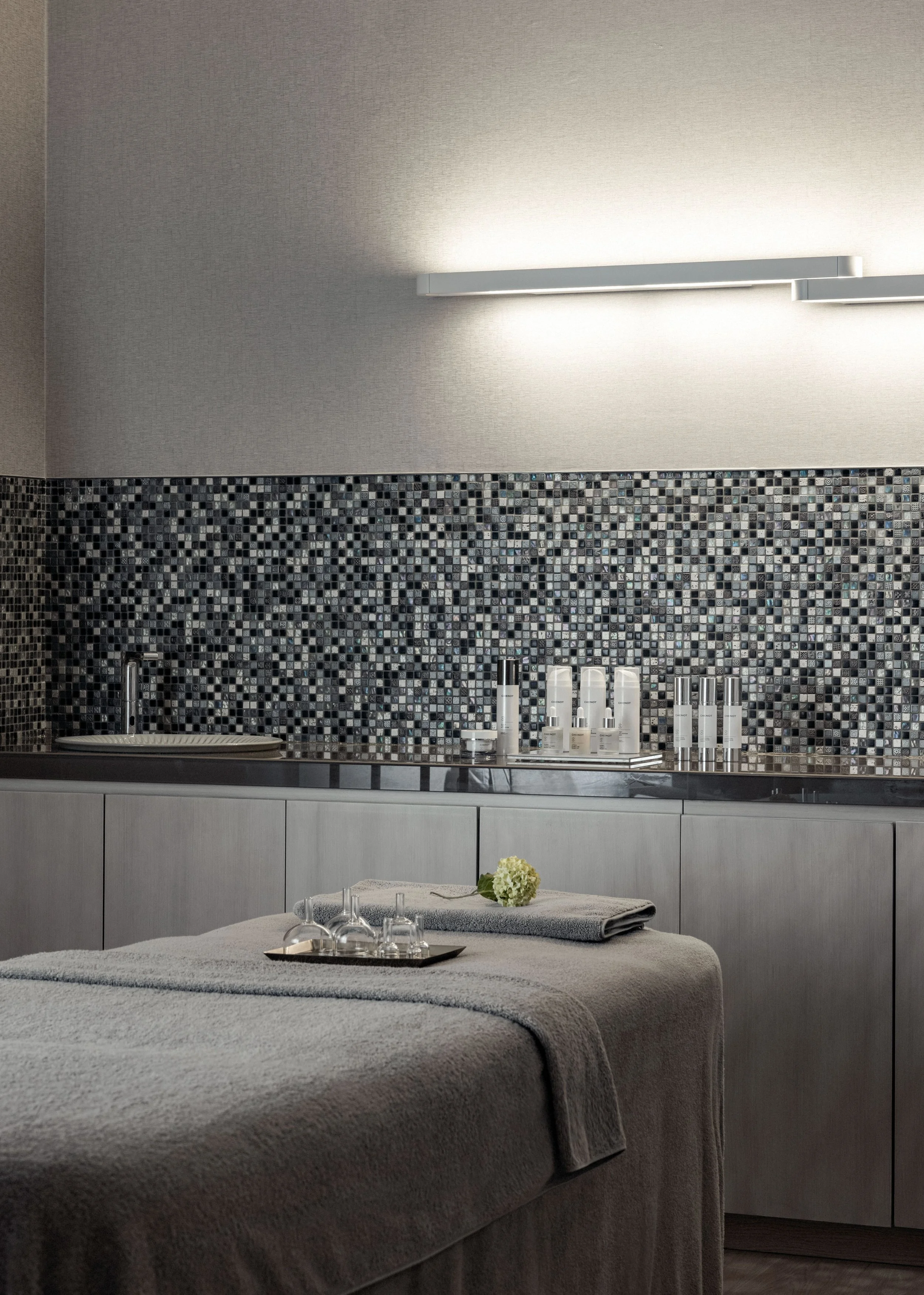 A spa or massage room with a bed covered in a gray towel, a tray with bottles and a towel, a flower, and a small table with glasses, a flower, and candles. The background has a tiled wall, a white cabinet, and a modern light fixture.
