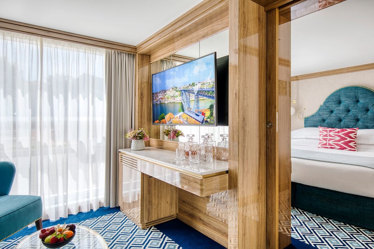 View of a hotel room with a bedroom on the right and a sitting area on the left. The bedroom has a teal upholstered headboard and a white bed with a pink patterned pillow. The sitting area features a flat-screen TV, a wooden and marble console table with floral arrangements and glass decanters, a blue armchair, and a round glass coffee table with a bowl of fruit. Floor-to-ceiling curtains cover large windows, and a blue patterned carpet covers the floor.
