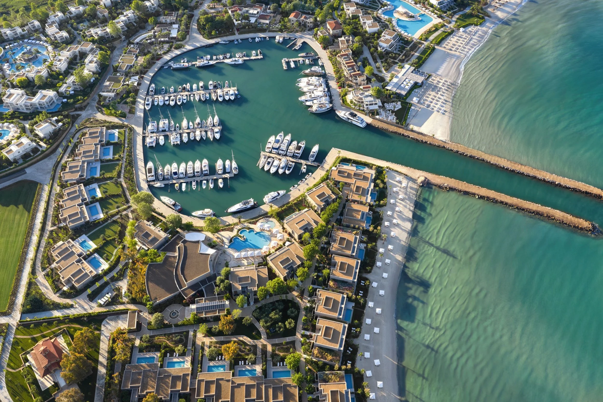 An aerial view of a coastal residential area with houses, swimming pools, and palm trees, a marina with boats and yachts, a sandy beach, and the ocean.