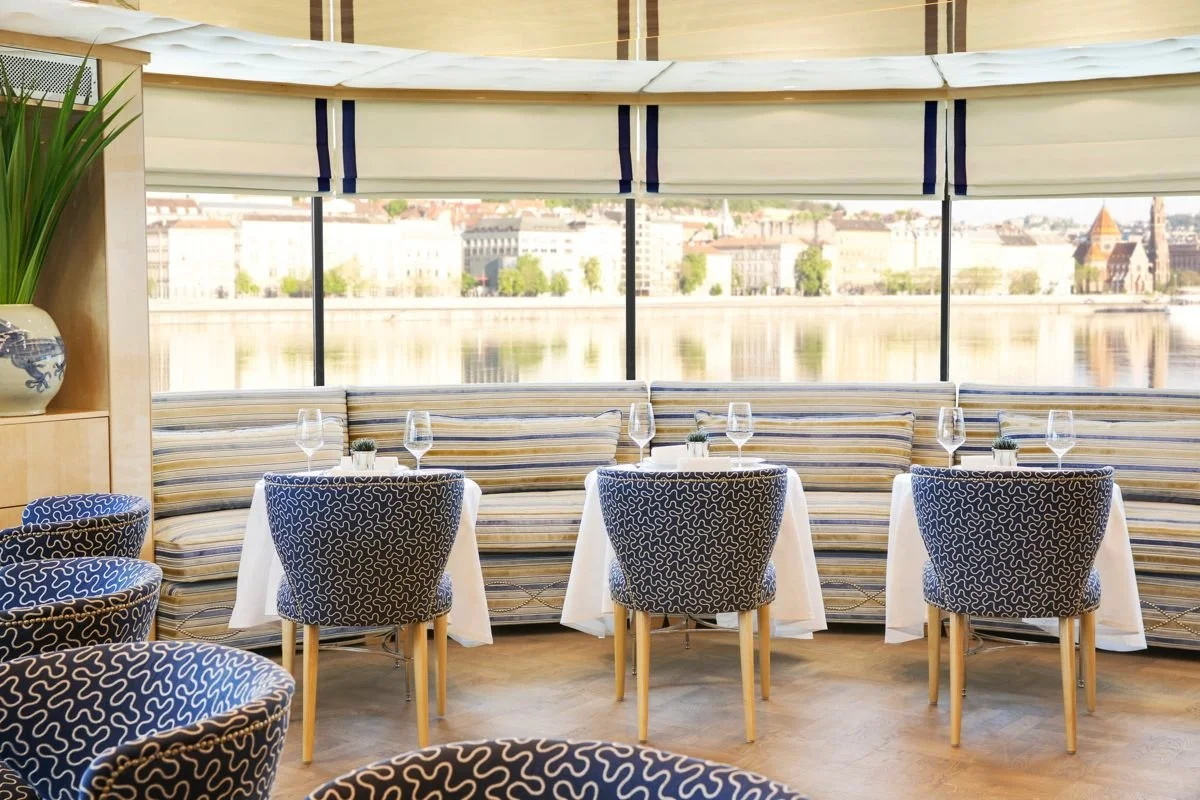 Elegant restaurant with white tablecloths, wine glasses, and patterned blue and white chairs, overlooking a river through large windows.