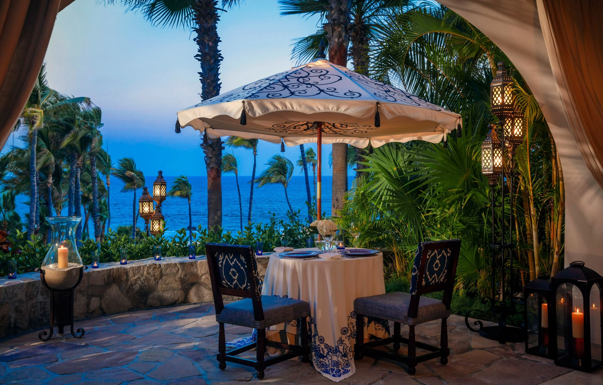 An outdoor dining area overlooking the ocean with a table set for dinner, decorated with candles, under a large ornate umbrella, surrounded by lush tropical plants and lanterns, during blue hour.