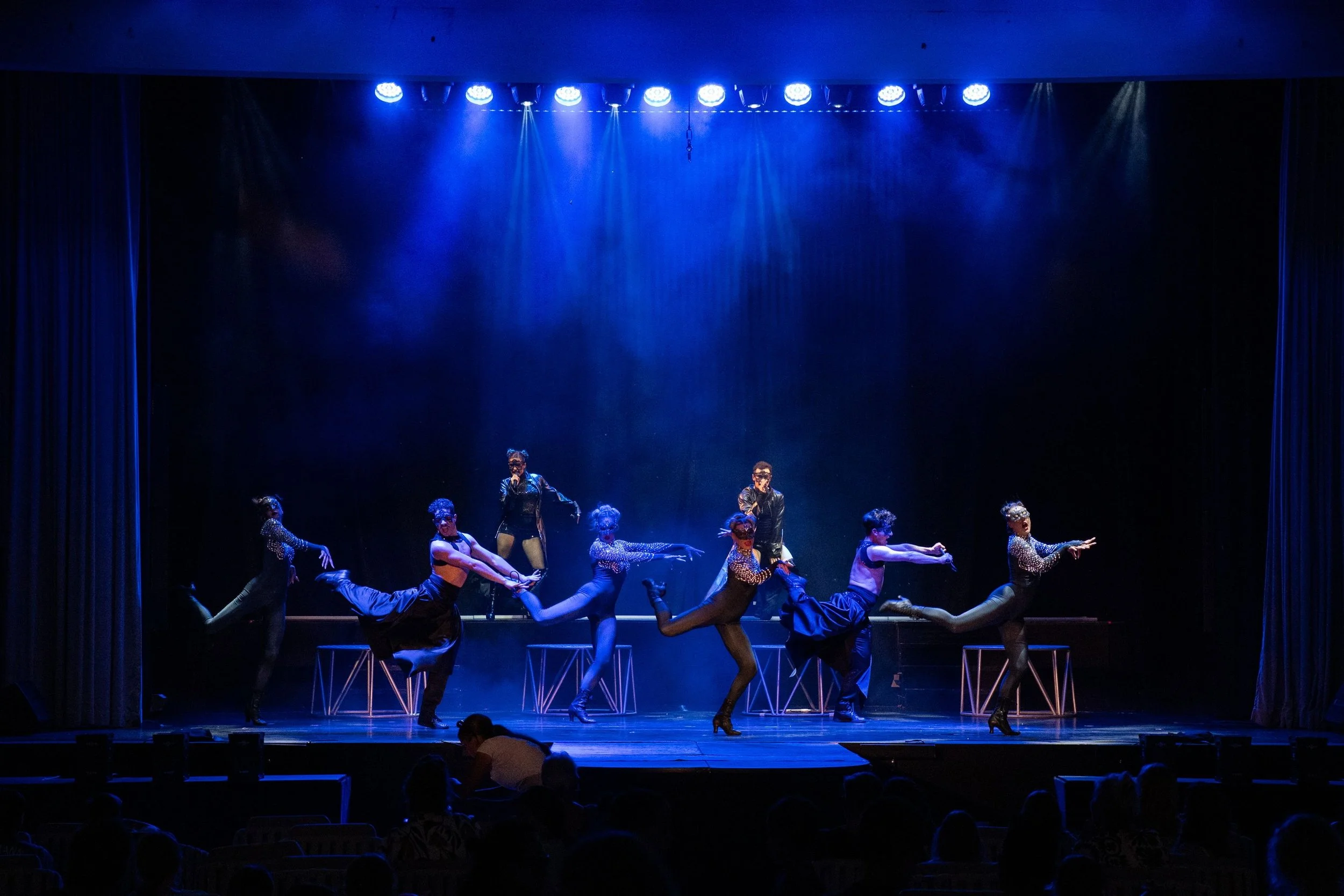 A stage performance with dancers in colorful costumes, some wearing polka dots and high heels, under blue stage lighting with a dark background.