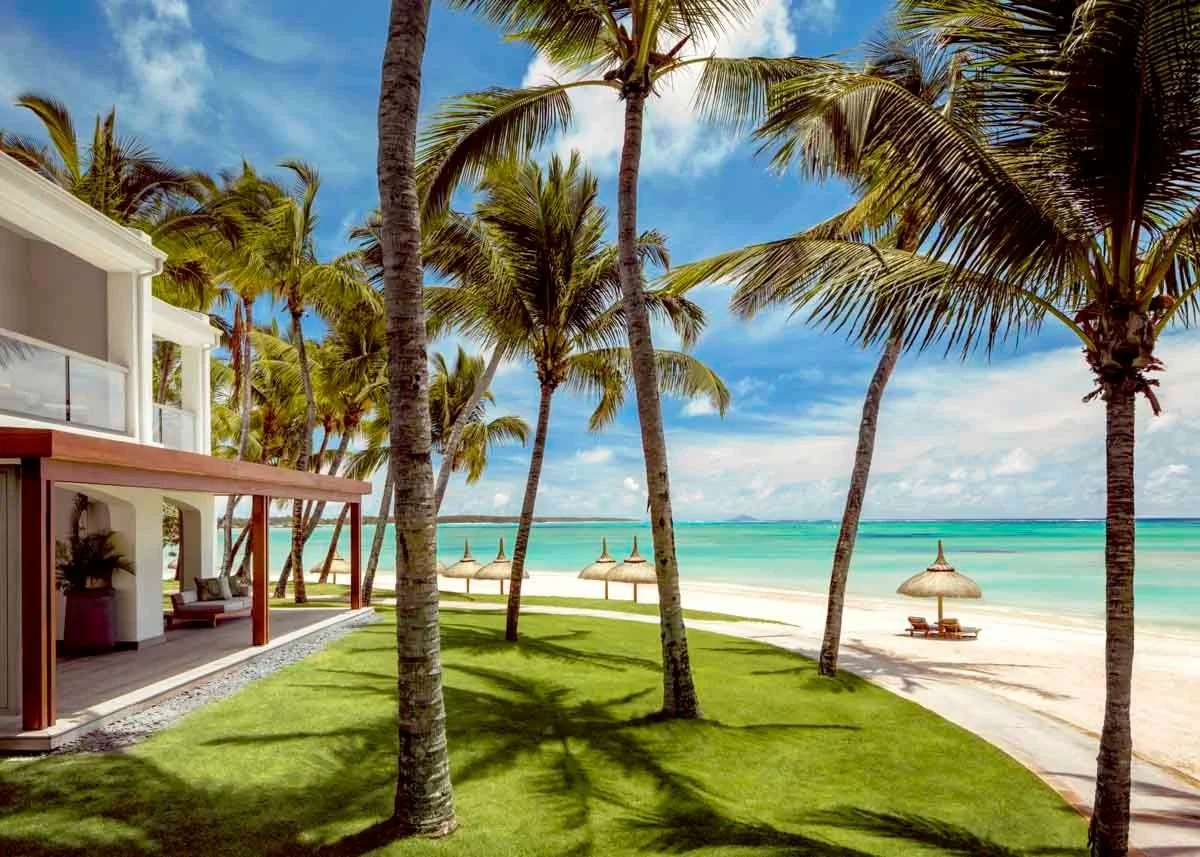 Tropical beach scene with tall palm trees, a grassy area, a sandy beach with straw umbrellas, lounge chairs, and calm turquoise ocean water under a partly cloudy sky.