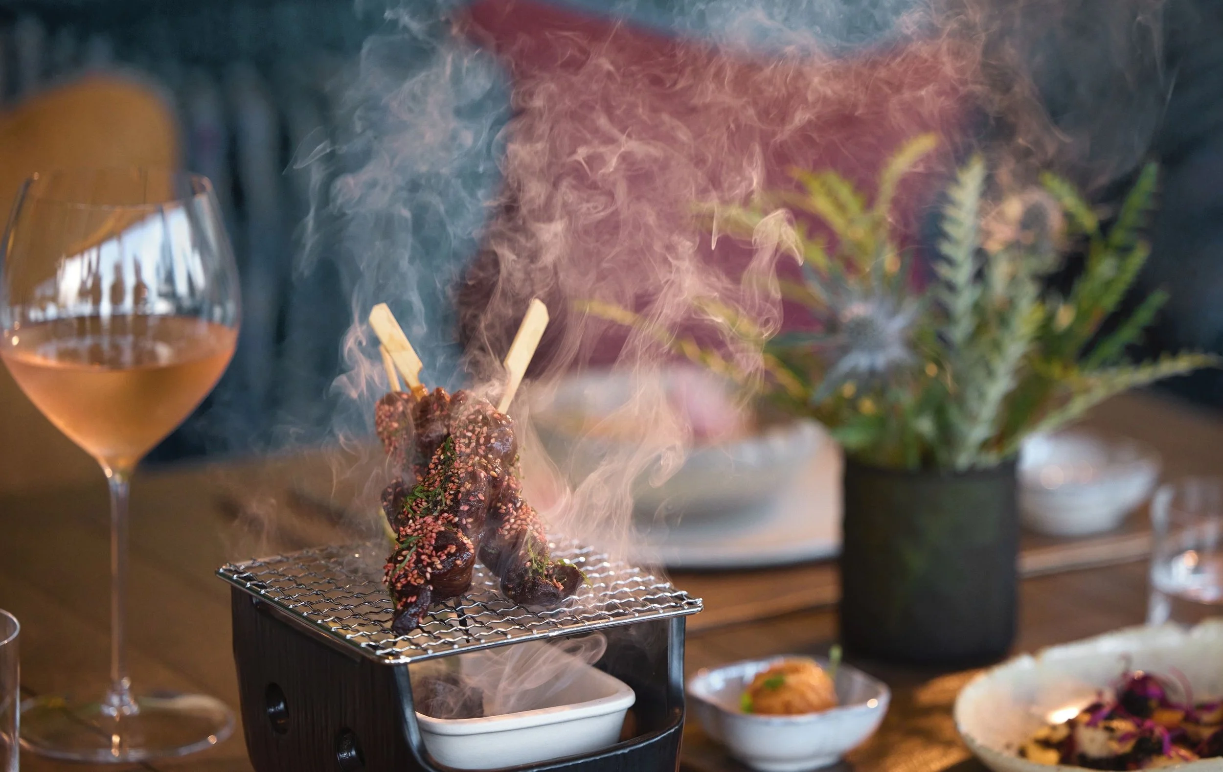 A dinner table with skewered grilled meat sprinkled with sesame seeds, with smoke rising from it, placed on a small grill. Next to it is a glass of rosé wine, and in the background, there are various dishes and a vase with flowers.