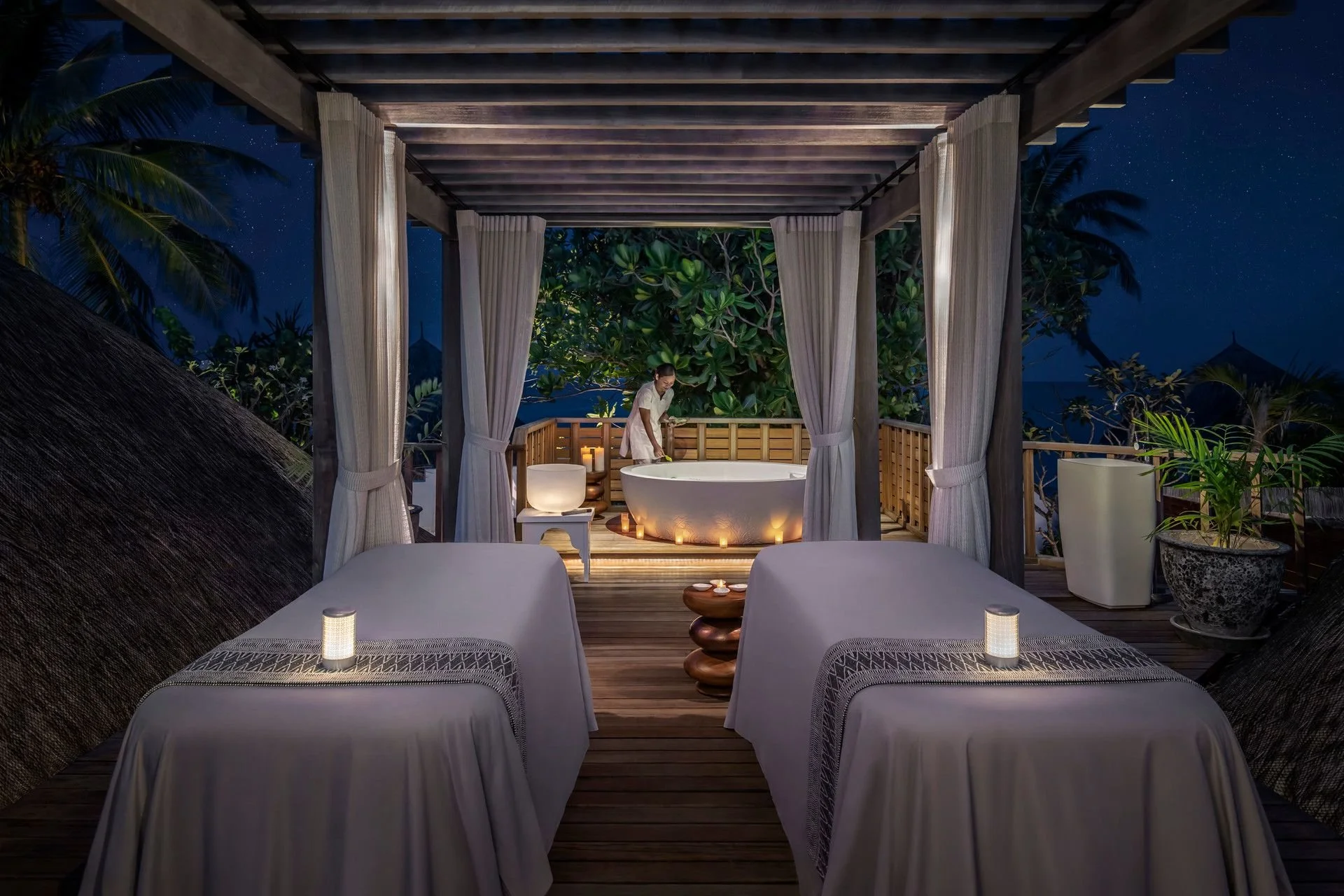 A woman relaxing in an outdoor bathtub on a wooden deck surrounded by greenery at night with a starry sky overhead.