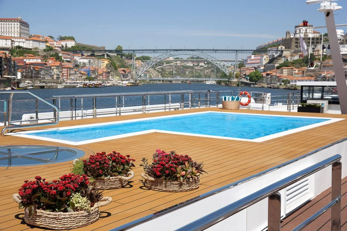 A view from the deck of a boat with a small swimming pool, potted flowers, and a basket on a river with a city skyline and bridge in the background.