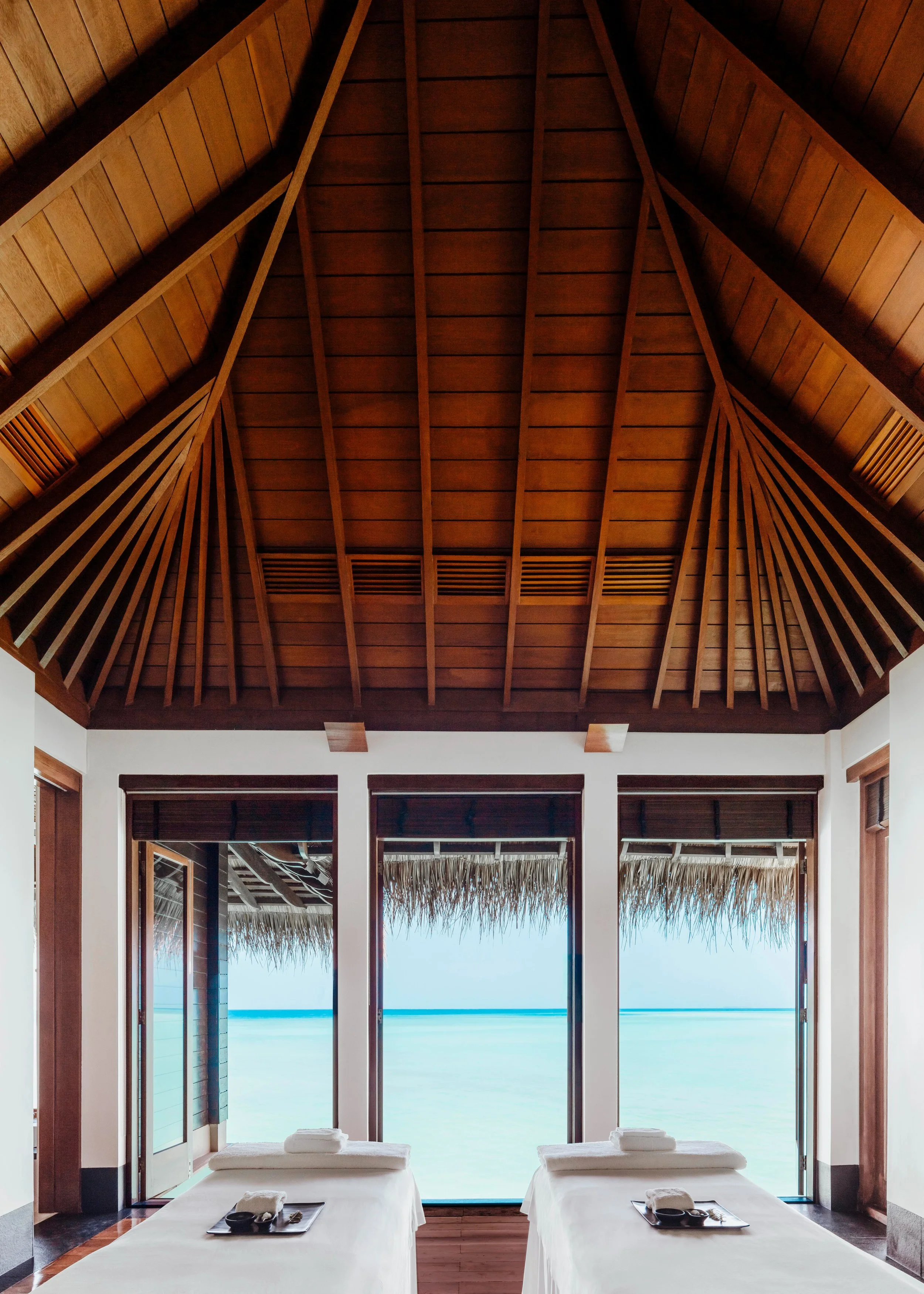 Two massage tables with white sheets and black trays, set indoors in front of large glass doors that overlook an ocean view with a clear sky.