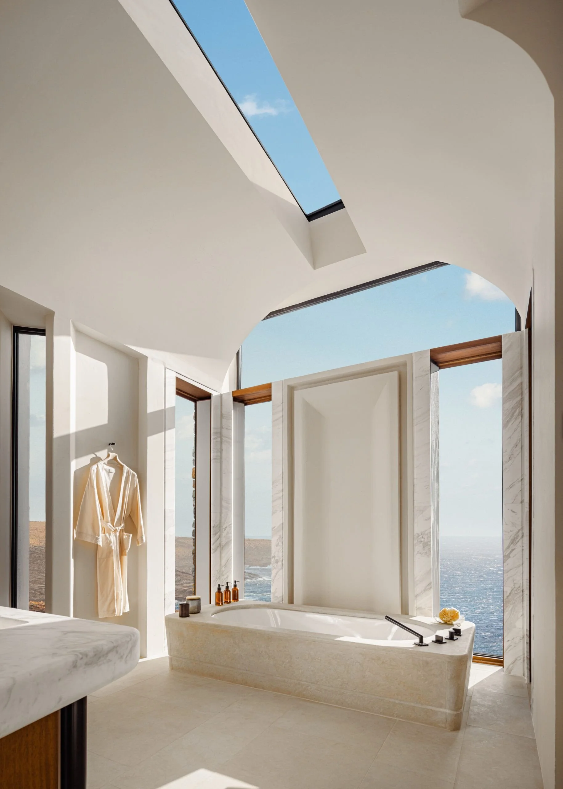 Modern bathroom with freestanding bathtub overlooking the ocean through large windows, with a white robe hanging and bathroom accessories on the counter.