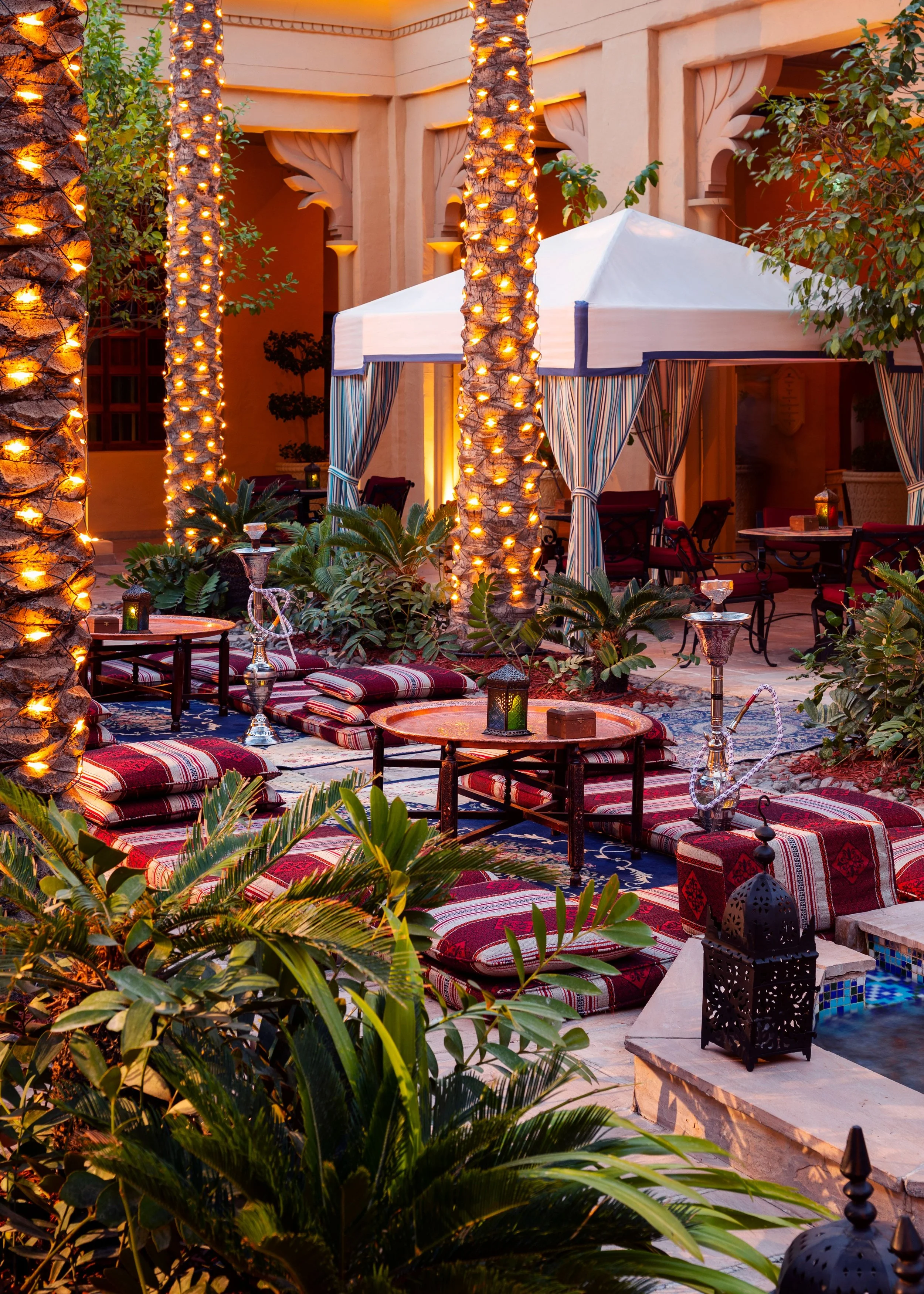 An outdoor patio with illuminated palm trees, colorful cushions on low tables, a white canopy tent with striped curtains, lush green plants, and lanterns, creating a cozy evening setting.
