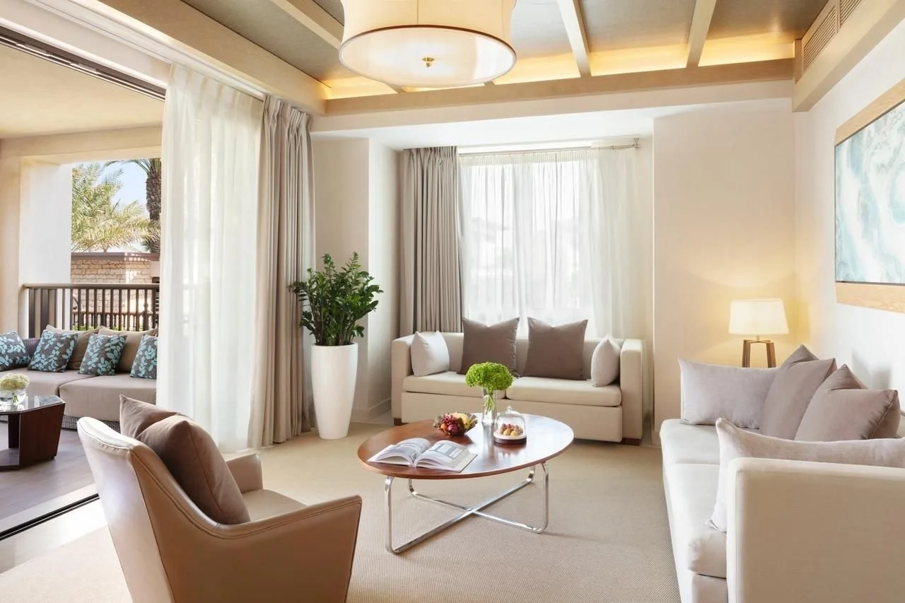 Bright living room with beige sofas, a wooden coffee table with flowers and an open book, large windows with curtains, a potted plant, and a balcony outside.