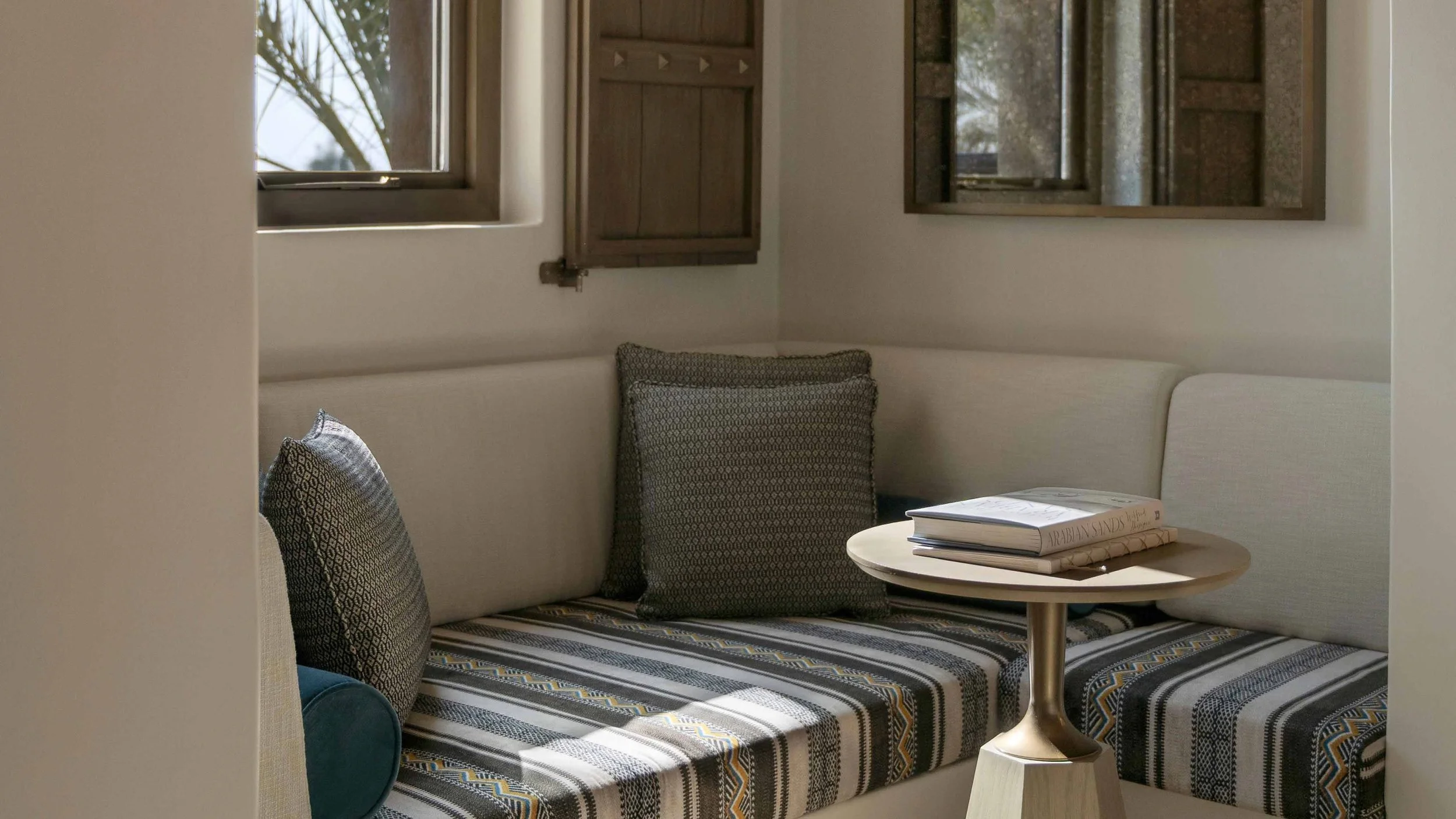 A cozy corner with a built-in beige cushioned bench with patterned pillows, a small round wooden table with magazines, and a window with wooden shutters.