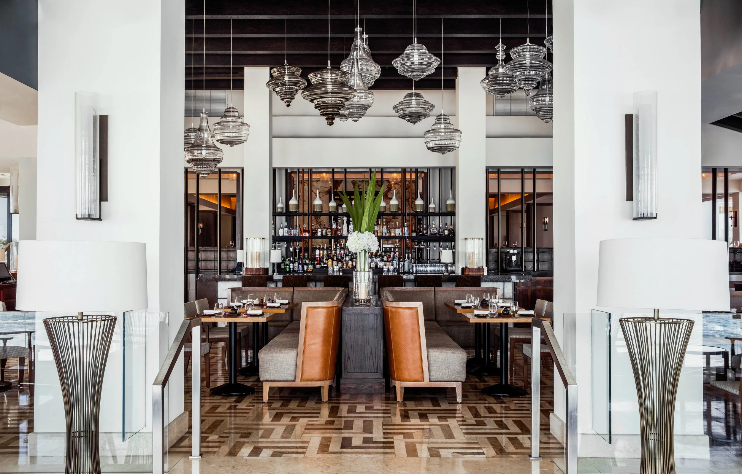Interior of a modern restaurant with a bar, glass tableware, a central flower arrangement, and contemporary lighting fixtures hanging from a dark wooden ceiling.