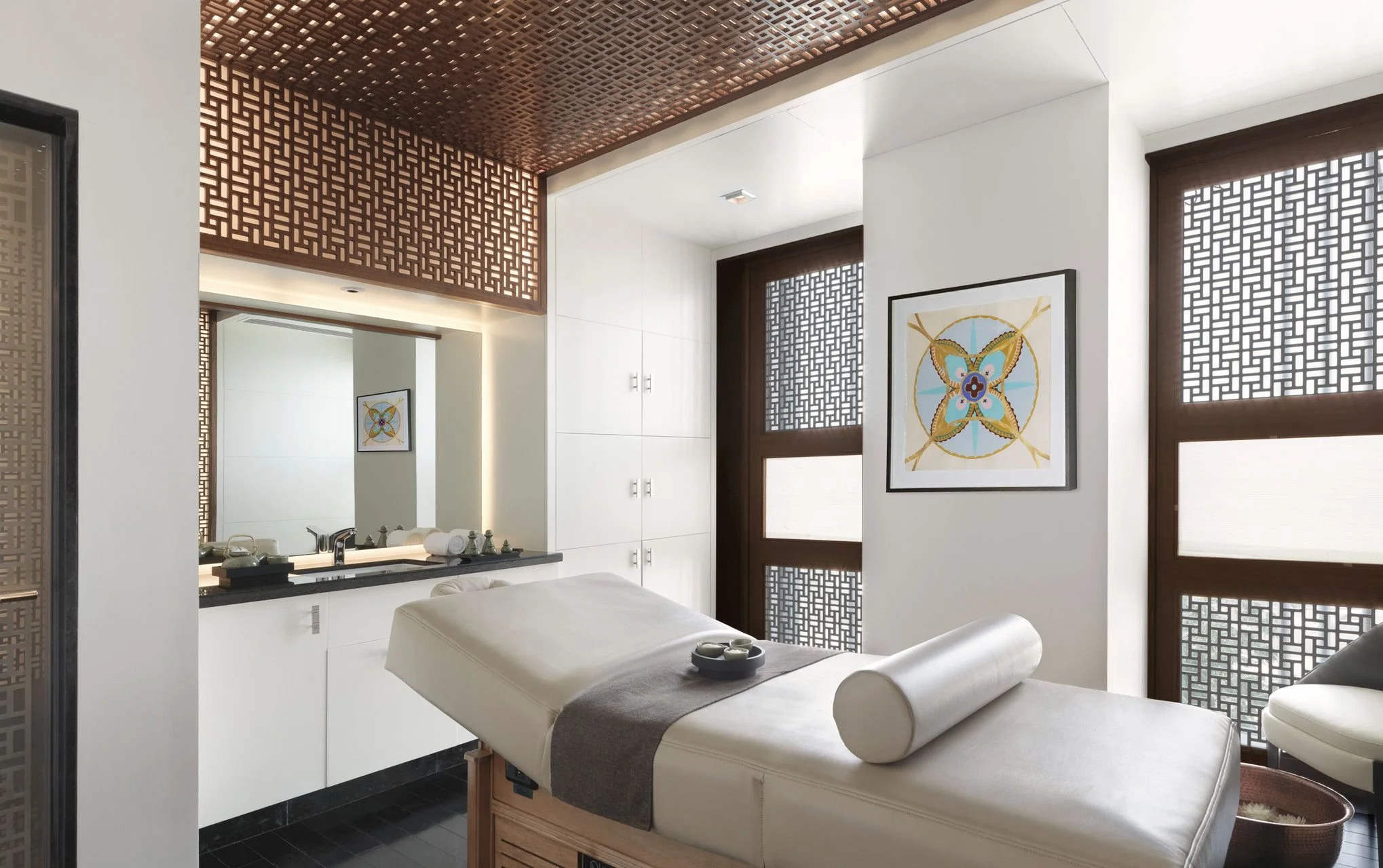 Modern spa room with white walls, wooden lattice windows, and a massage table with white sheets and a rolled towel. There are two framed artworks on the wall featuring colorful floral patterns. A mirror and a countertop with toiletries are visible in the background.