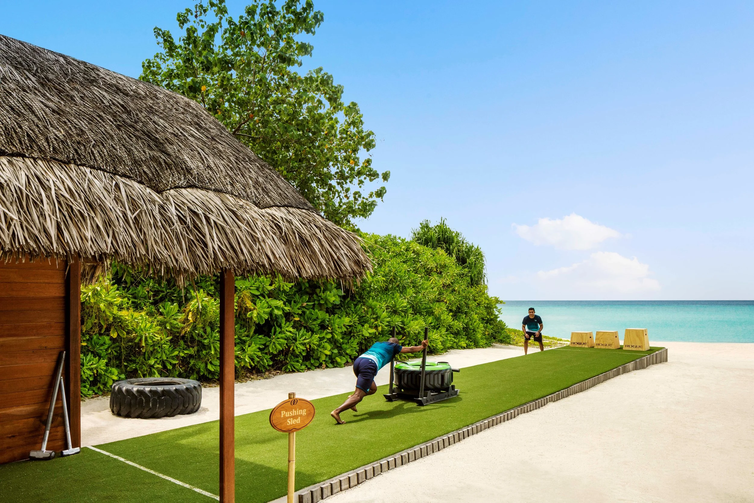 Two men are outdoors on a lawn, one pushing a sled labeled 'Pushing Sled' and the other preparing to lift a heavy tire. They are near a thatched-roof structure, lush green bushes, and a sandy beach with the ocean in the background. The sky is mostly clear with a few clouds.