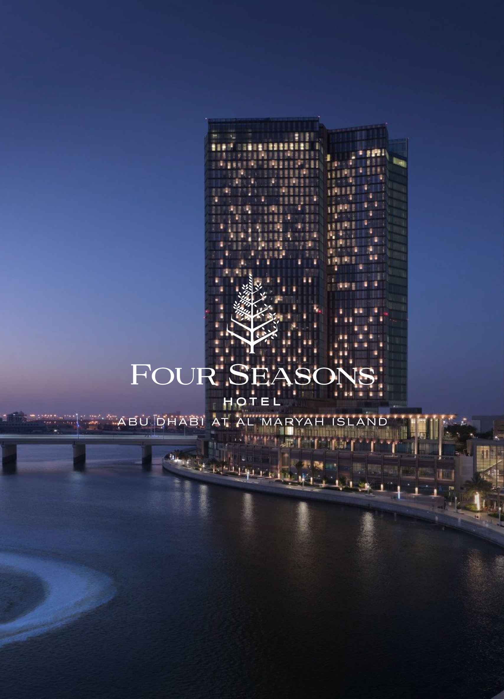 Night view of a modern high-rise hotel building with illuminated windows, situated along a river with a bridge, at Al Maryah Island in Abu Dhabi.