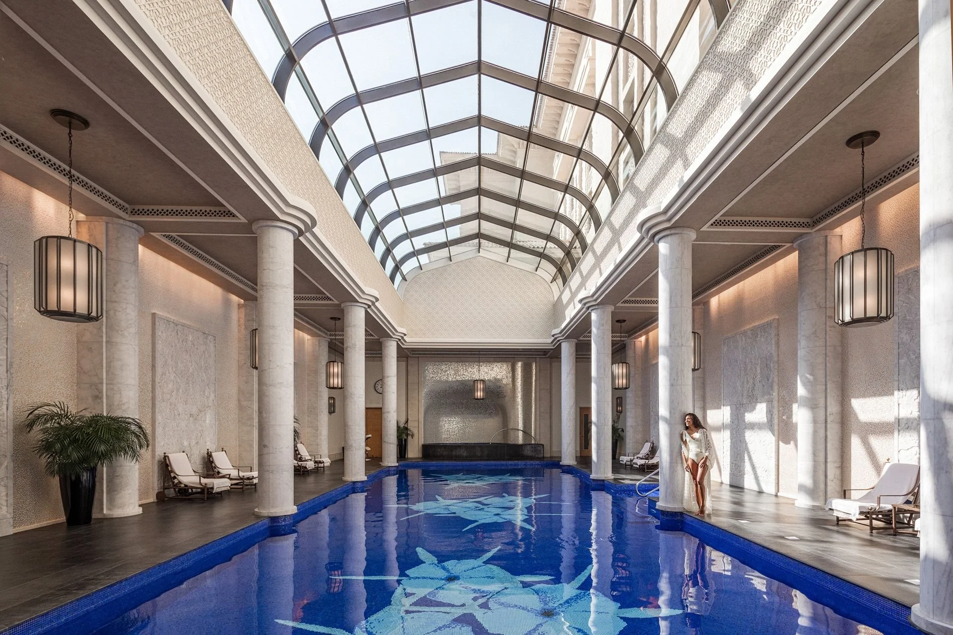 Indoor swimming pool area with white columns, a glass ceiling, lounge chairs, and a woman standing near the pool.