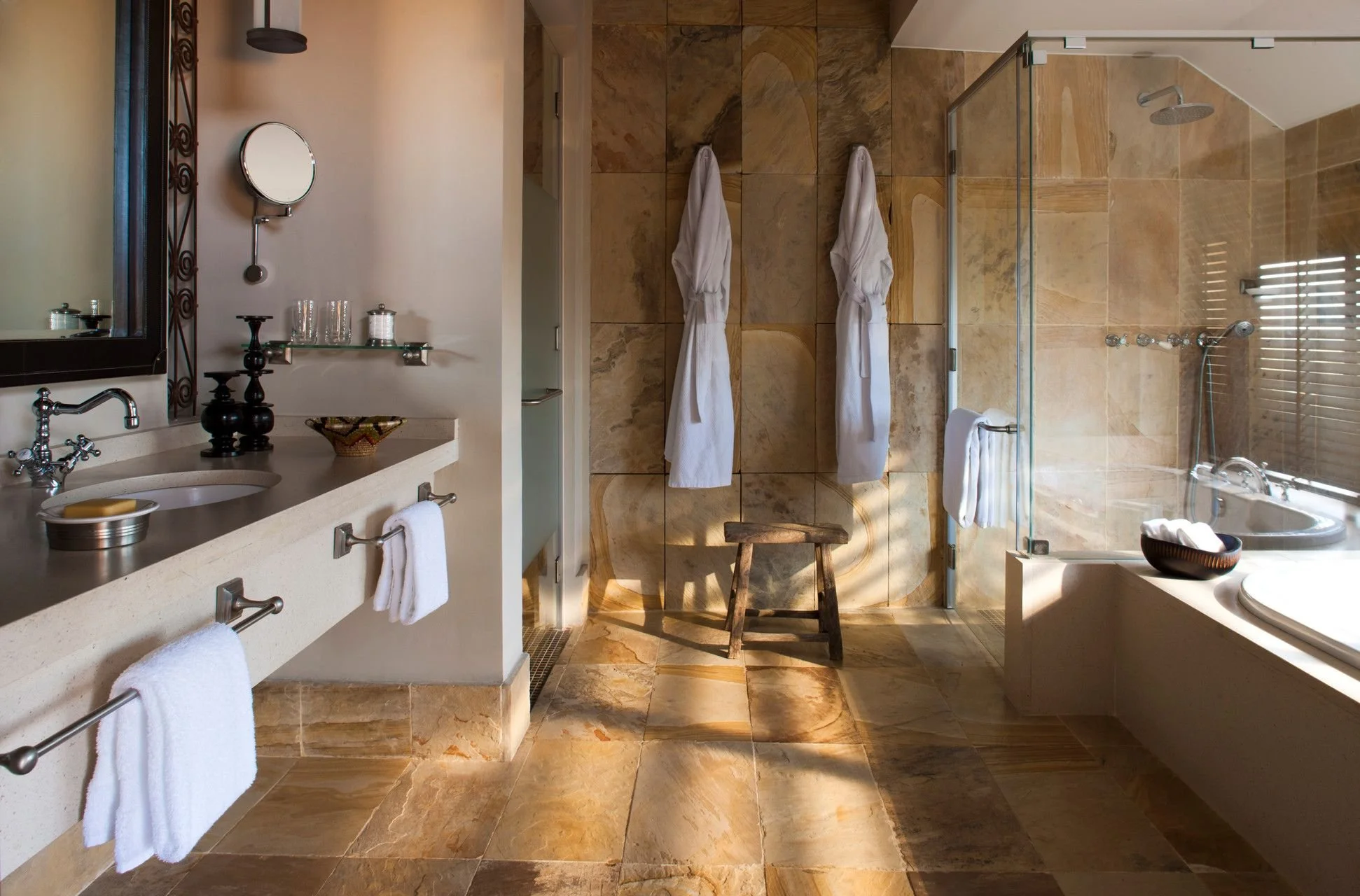 A luxurious bathroom with beige marble tiles on the floor and wall, a double sink vanity with towels hanging on racks, and a large walk-in shower with glass doors and a rain shower head, decorated with robes hanging on the wall and a small stool.
