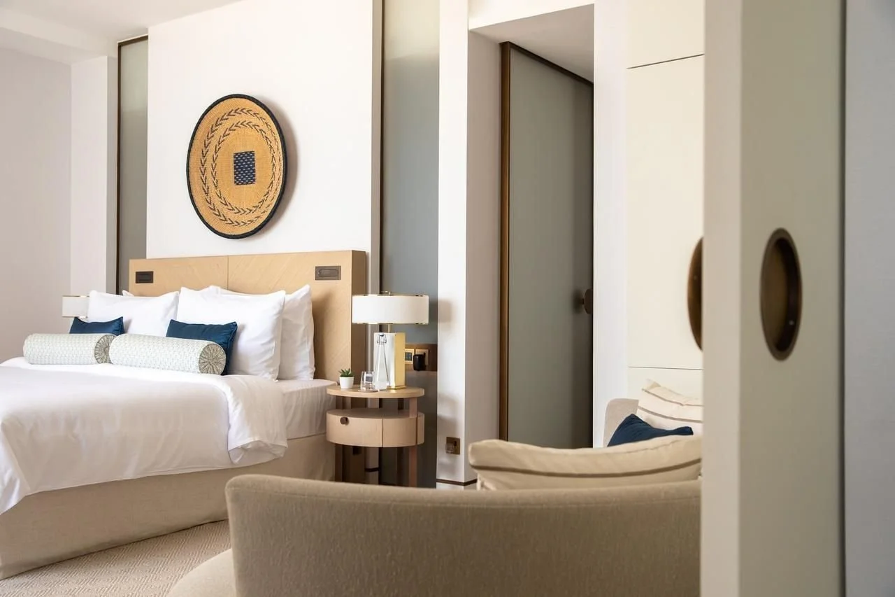 Modern hotel room with a white bed, blue pillows, a round side table with a lamp, and a beige armchair. A decorative circular wall art hangs above the bed. The room has a neutral color palette with light walls and minimal decor.