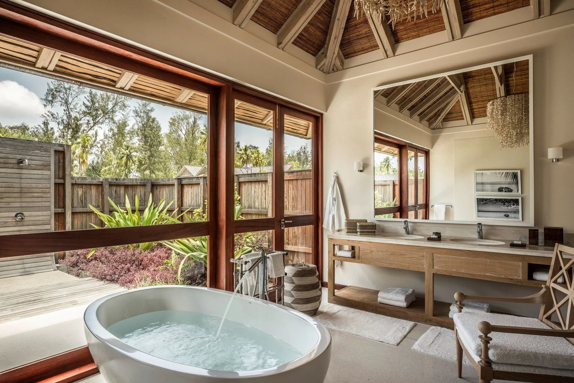 Bathroom with a large window overlooking a fenced backyard garden, featuring a freestanding bathtub, wood vanity with a sink, and decorative items.
