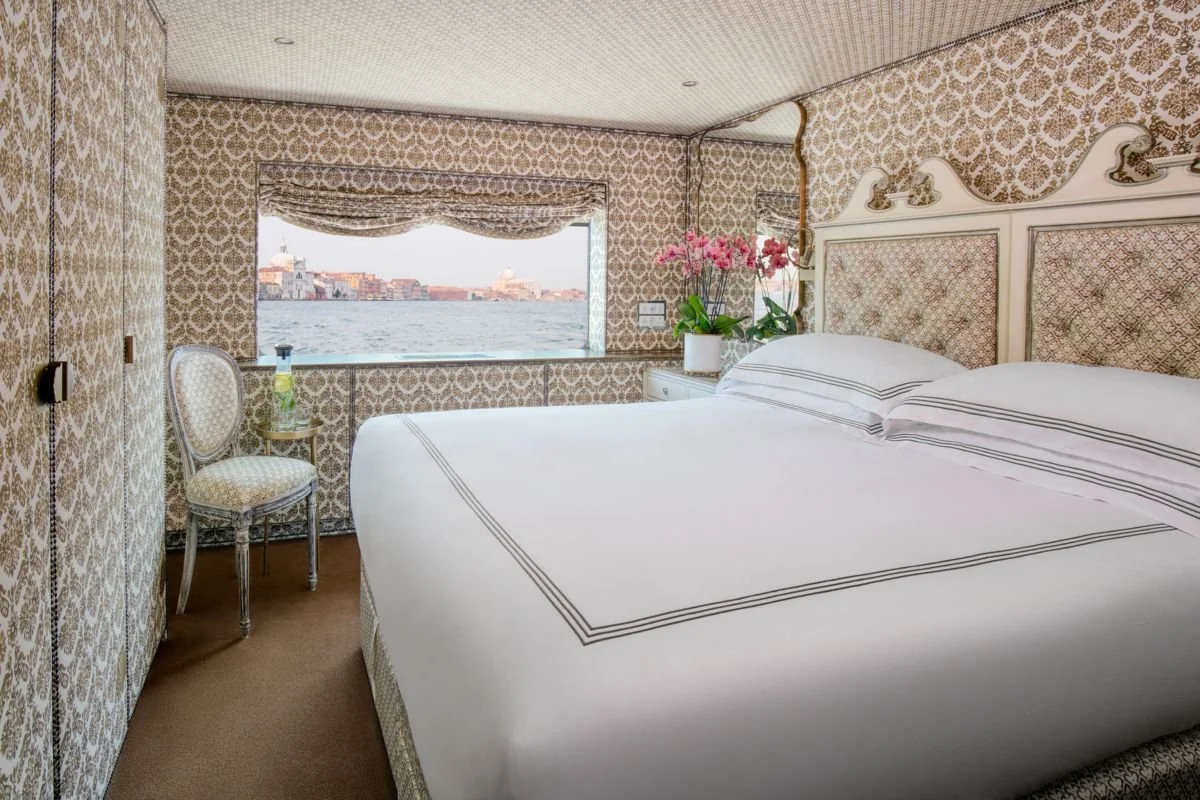 A hotel room with a large bed, floral wallpaper, a window with a view of a body of water and buildings, a chair, and a lamp with pink flowers on the nightstand.
