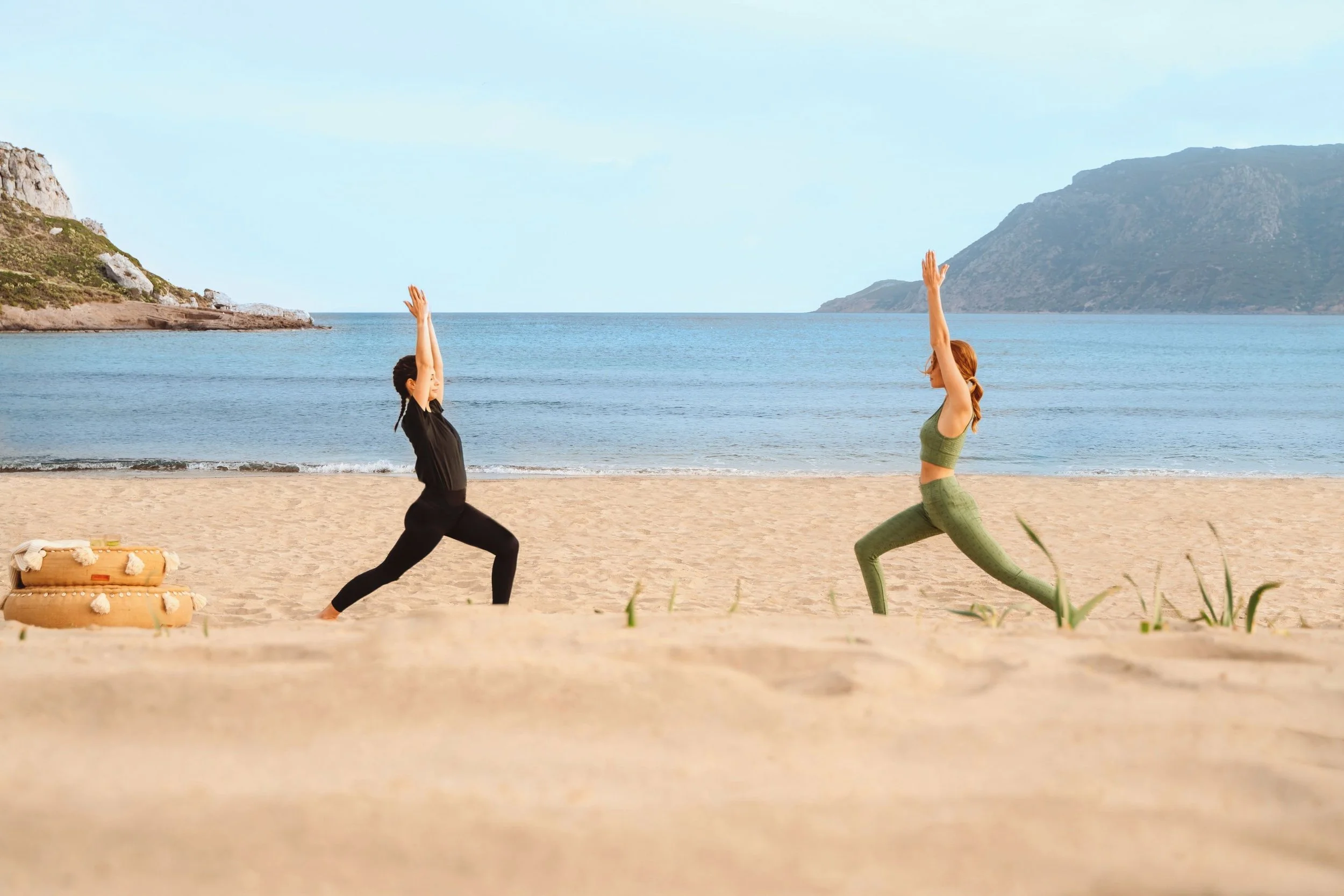 Two women practicing yoga on a sandy beach near the ocean, with mountains in the background and a small watermelon-shaped organizer on the sand.