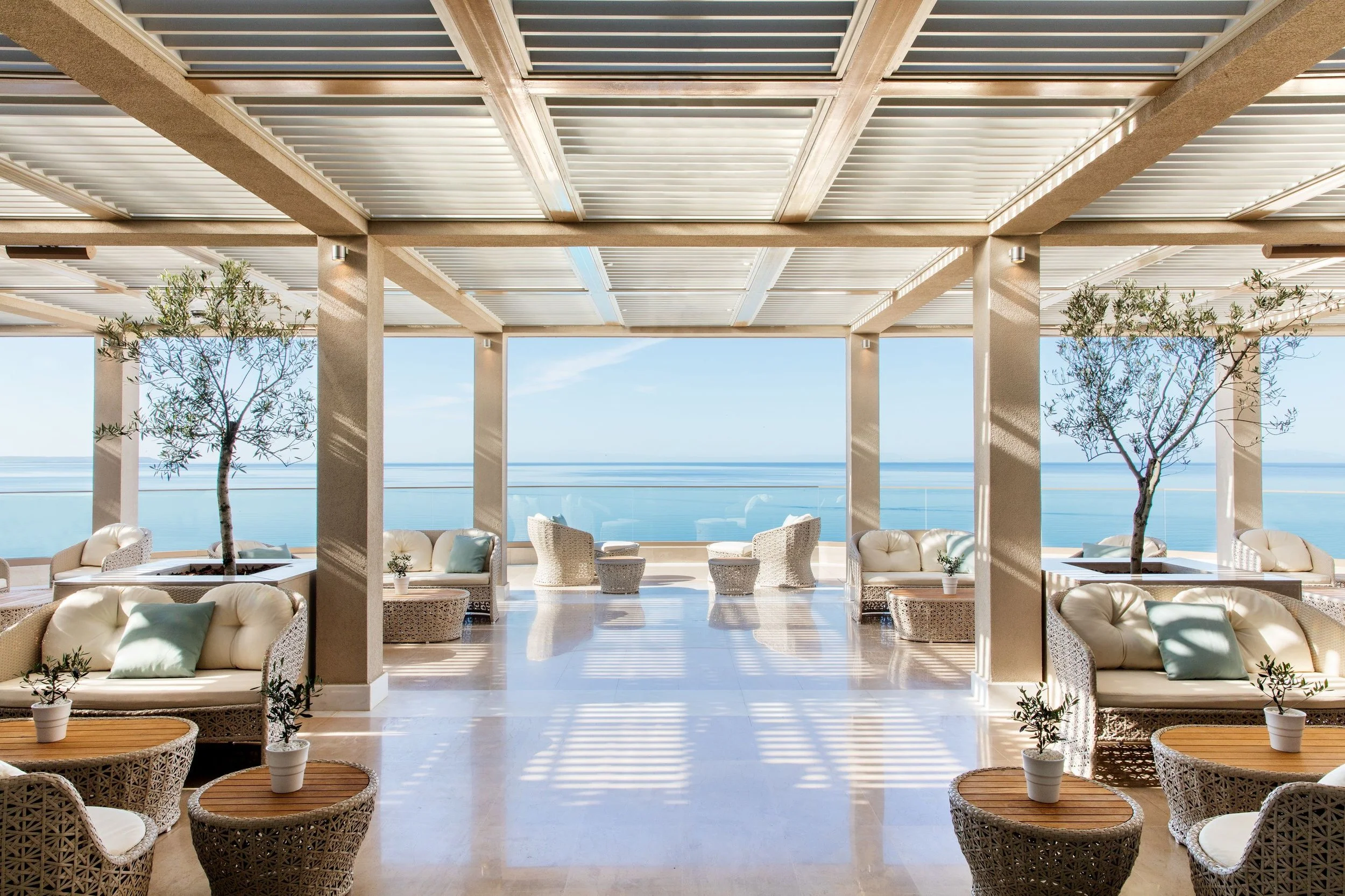 Outdoor lounge area with wicker chairs, potted plants, and a view of the ocean under a wooden pergola.