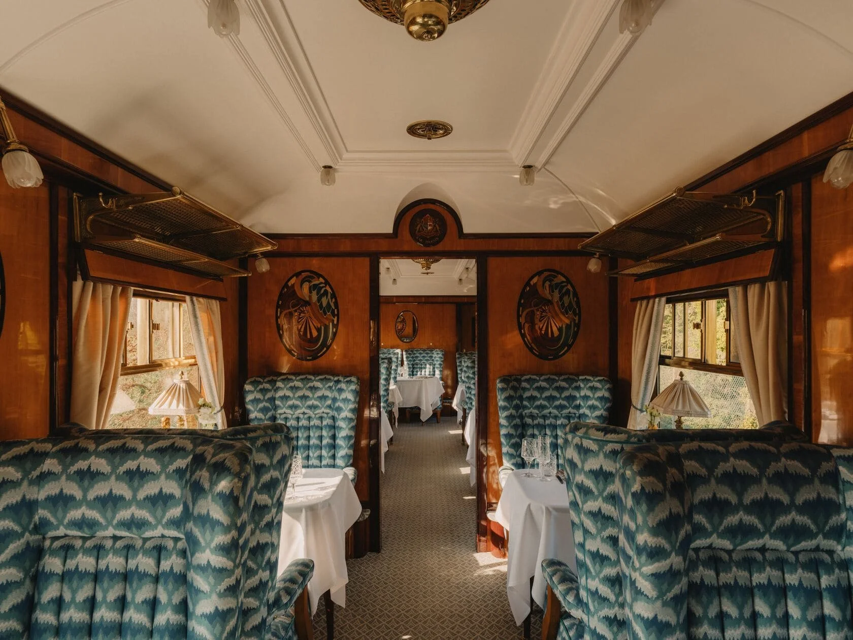 Interior of a vintage train dining car with blue patterned upholstery, white tablecloths, windows with curtains, and decorative wood paneling.