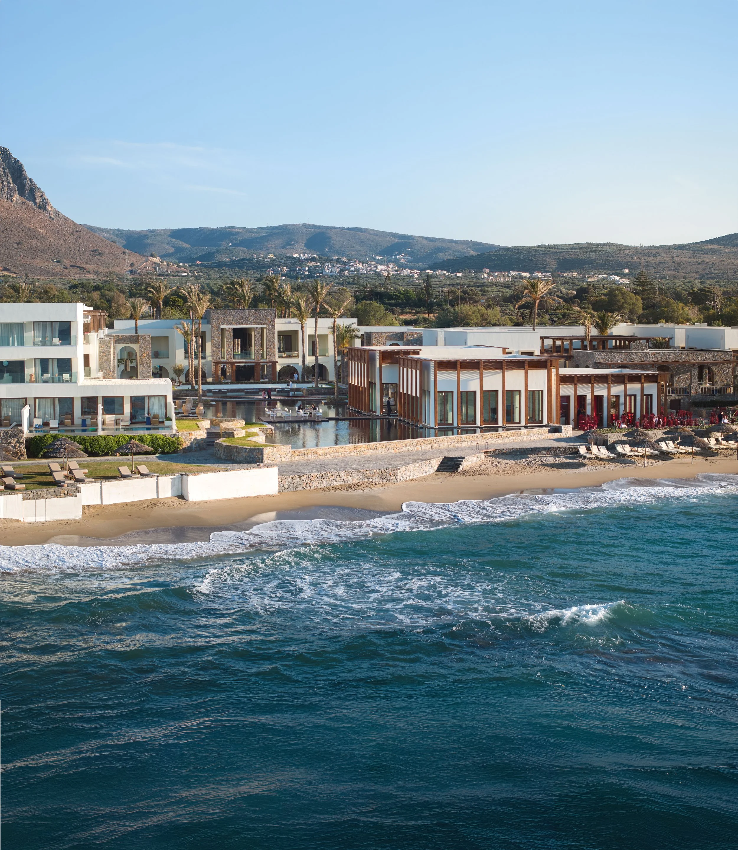 View of a resort with a sandy beach, lounge chairs, a pool, and modern white buildings with wooden accents, overlooking the ocean with waves.