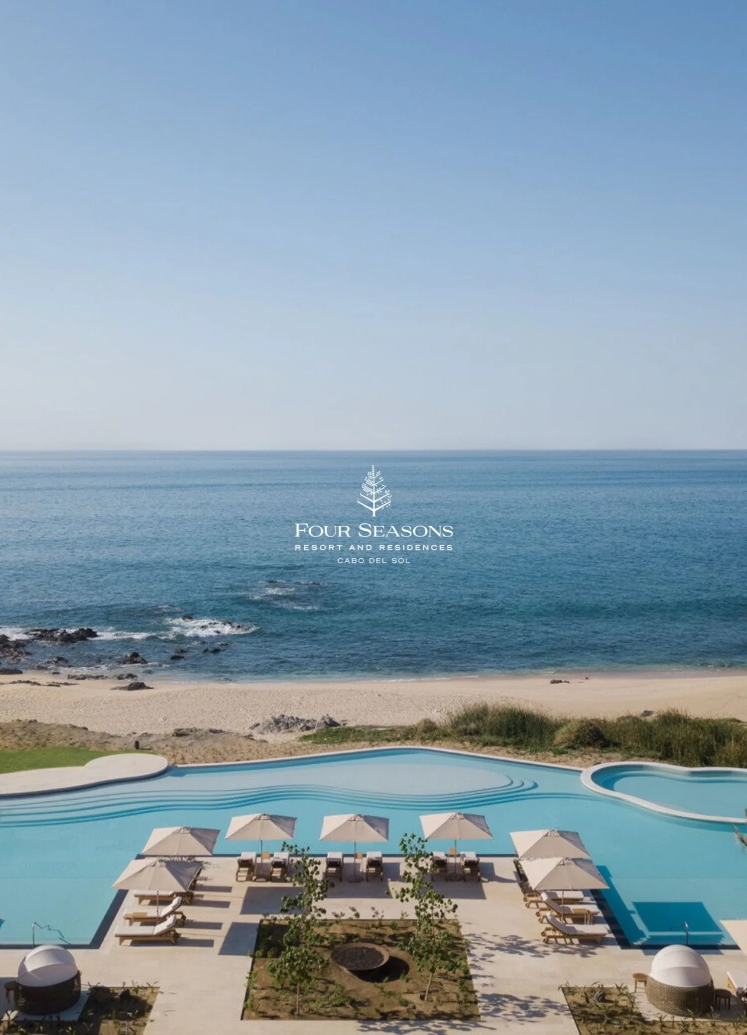 A coastal resort with a swimming pool and sun umbrellas, overlooking the ocean and sandy beach with rocks, under a clear blue sky.