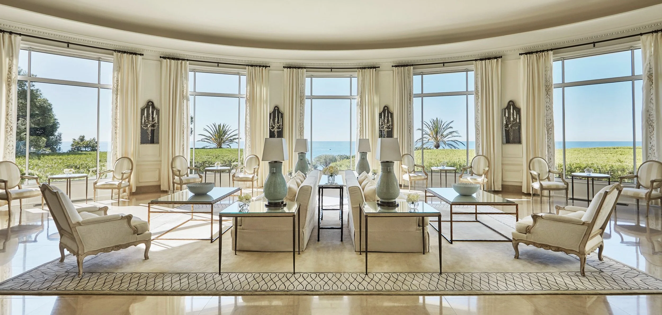 Luxurious living room with large floor-to-ceiling windows showing an ocean view, white curtains, elegant furniture, and decorative lamps.