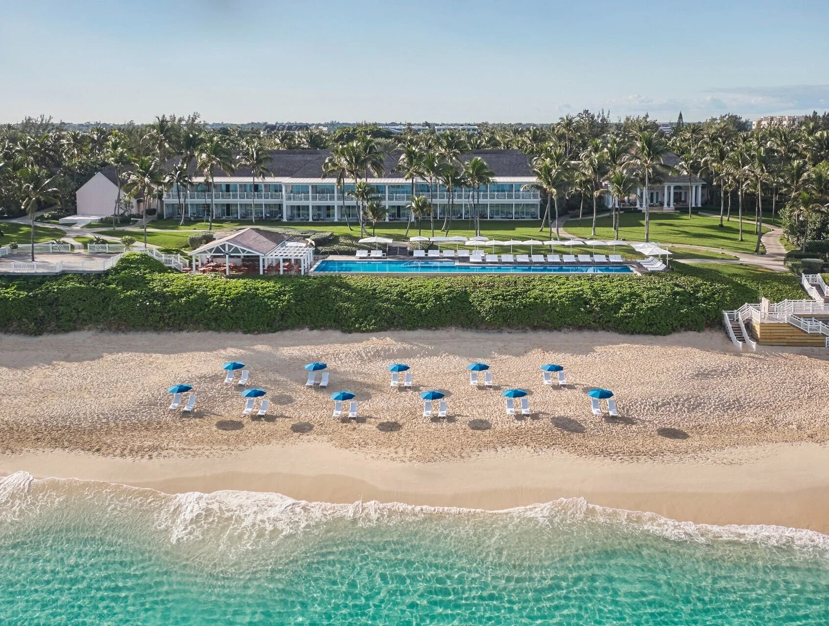 Beach with ten blue umbrellas and lounge chairs, ocean waves, grassy area, swimming pool, palm trees, and a large white building in the background.