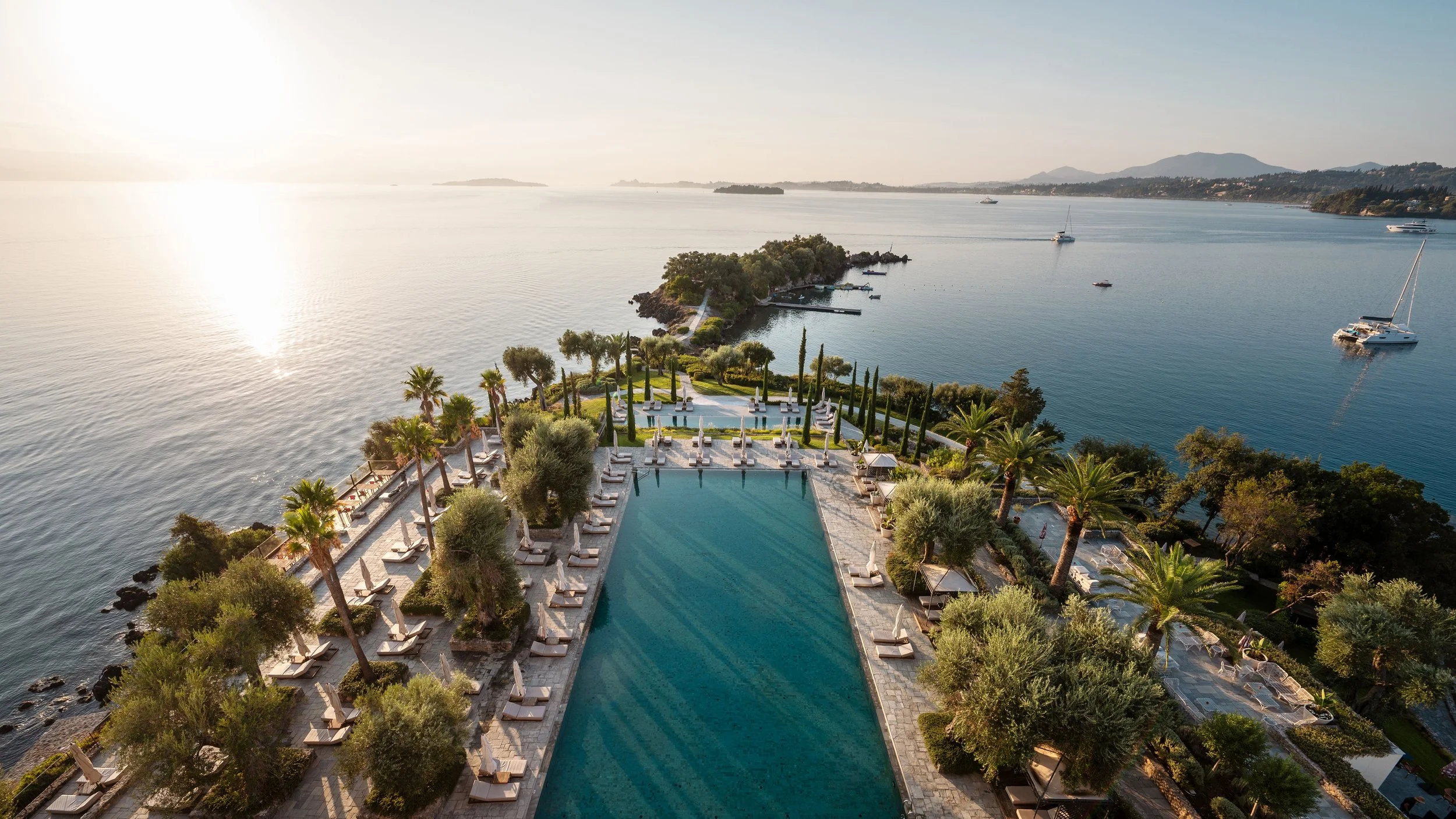 Aerial view of a luxurious infinity pool overlooking a large body of water with boats and islands, surrounded by palm trees and sun loungers, during sunset.